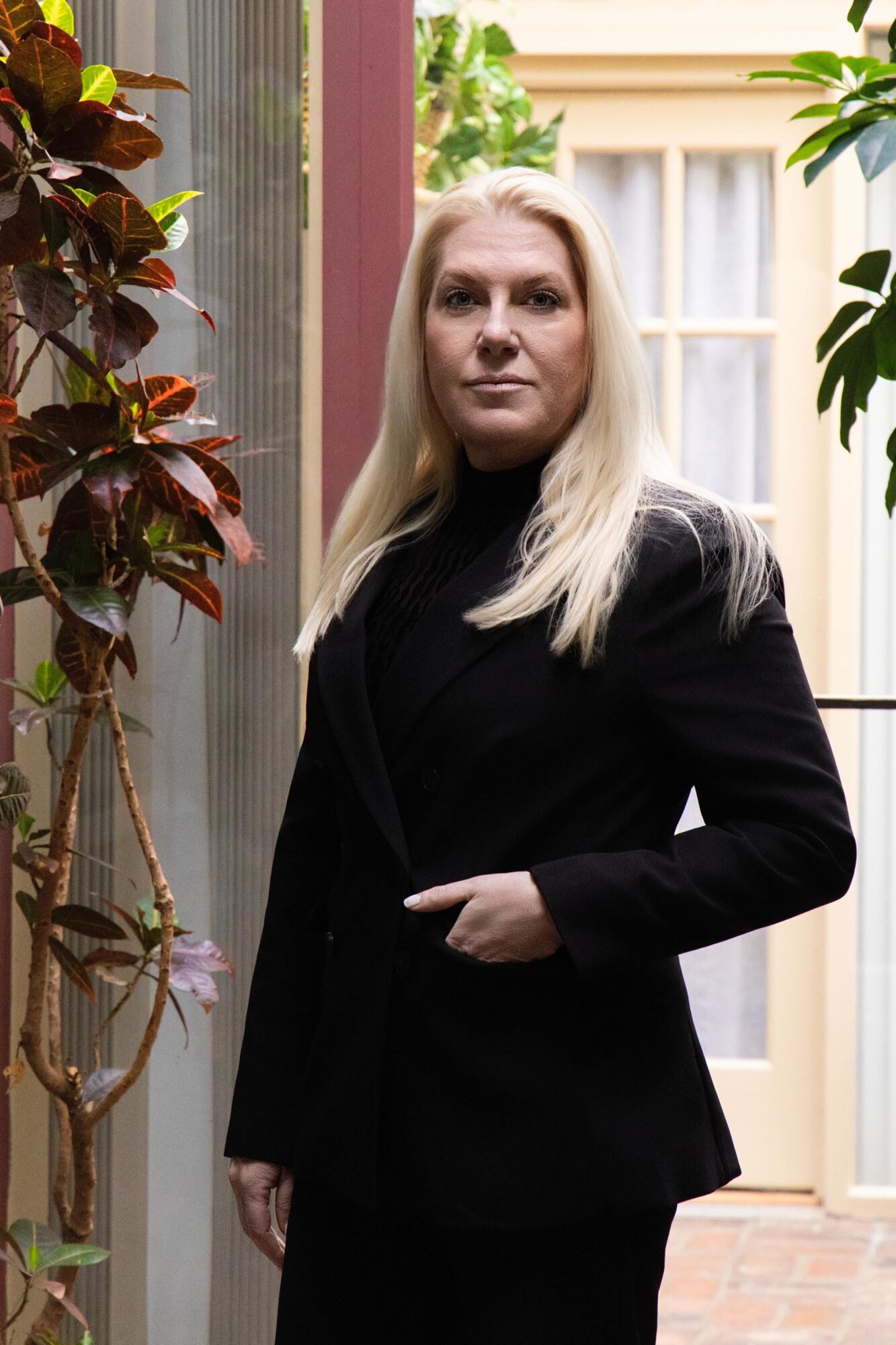 A person with long blonde hair stands indoors in black clothing beside leafy plants and a window.