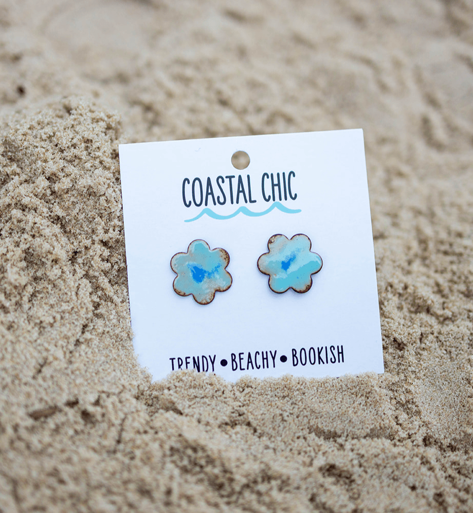 White card labeled “Coastal Chic” with two blue flower-shaped earrings resting in sand.
