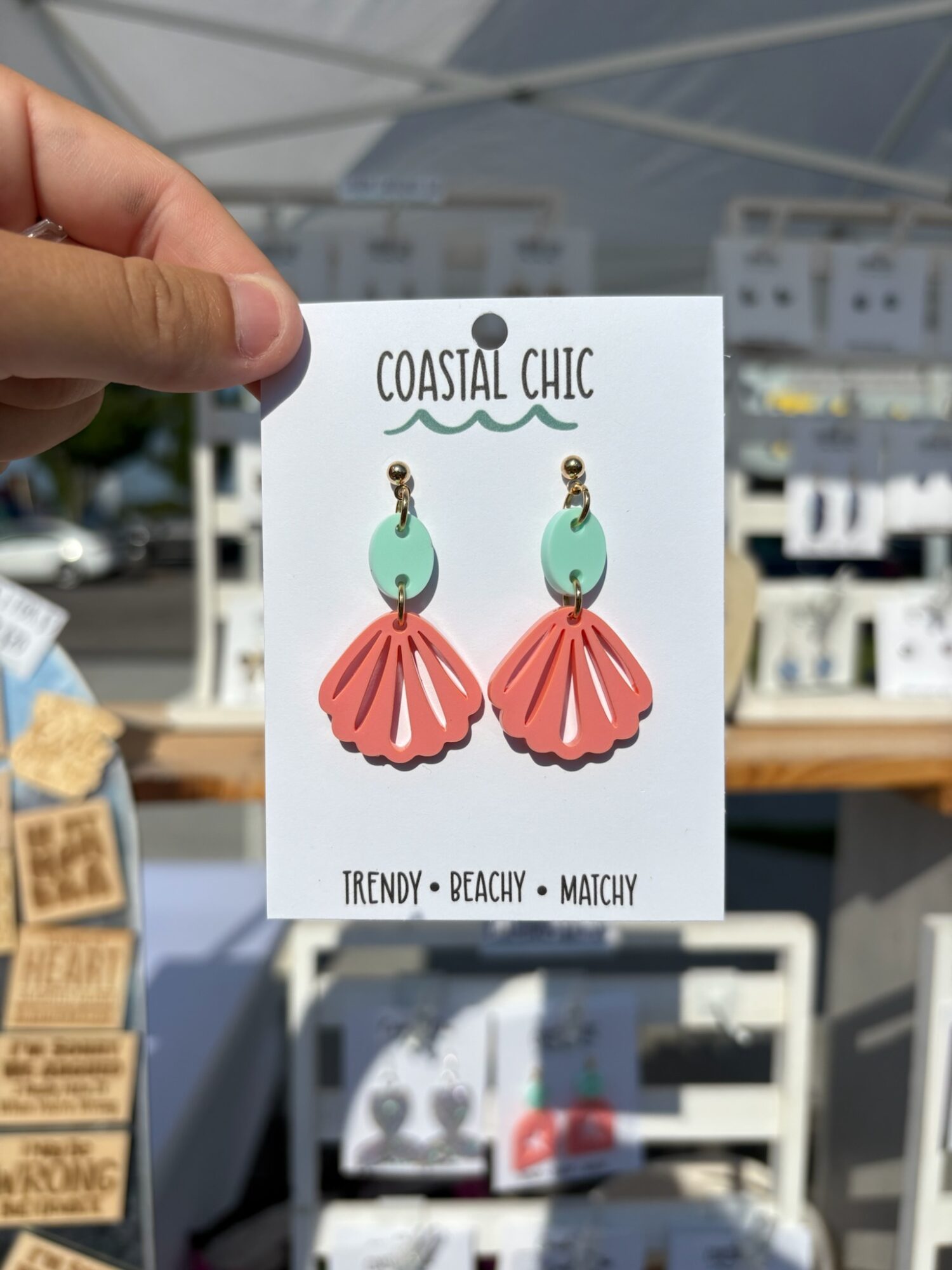 A hand holds a card displaying two dangling earrings with green stones and coral-colored fan-shaped pieces.