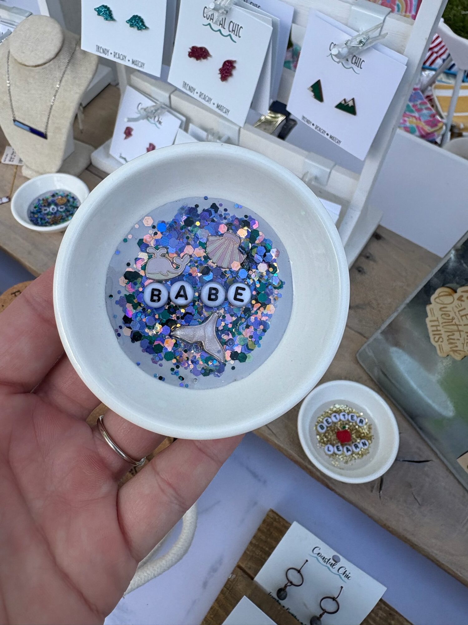 A hand holds a white dish filled with colorful glitter and letter beads reading “BABE,” among jewelry displays.