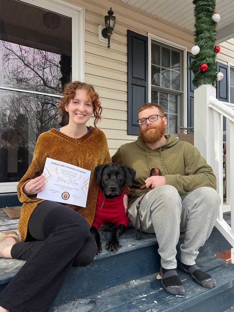 Two people sit on porch steps with a black dog wearing a red sweater; one person holds a certificate.