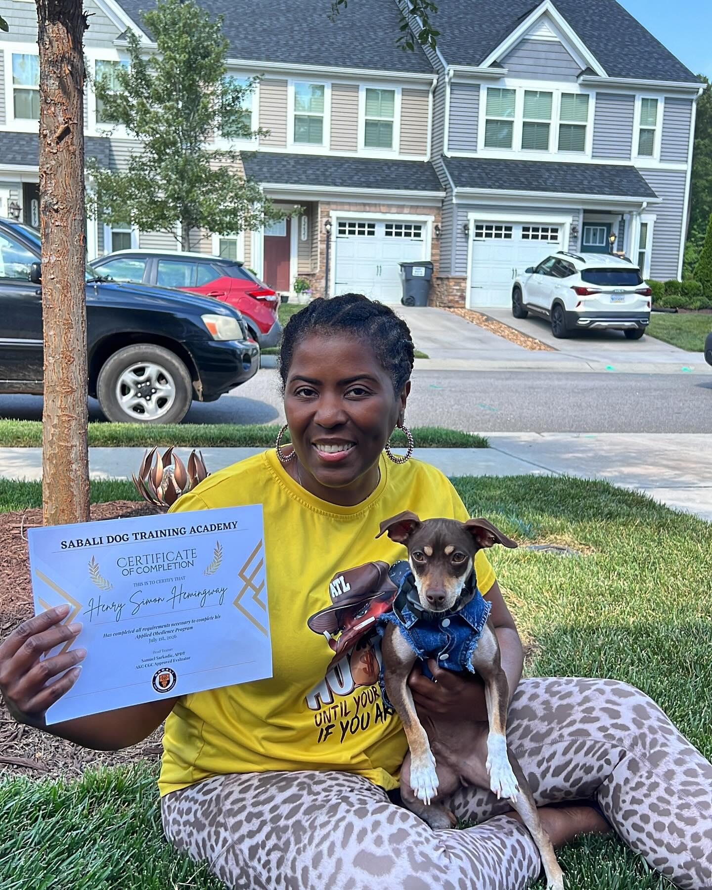 Person holding a certificate sits on grass with a small dog in their lap and a larger dog beside them.