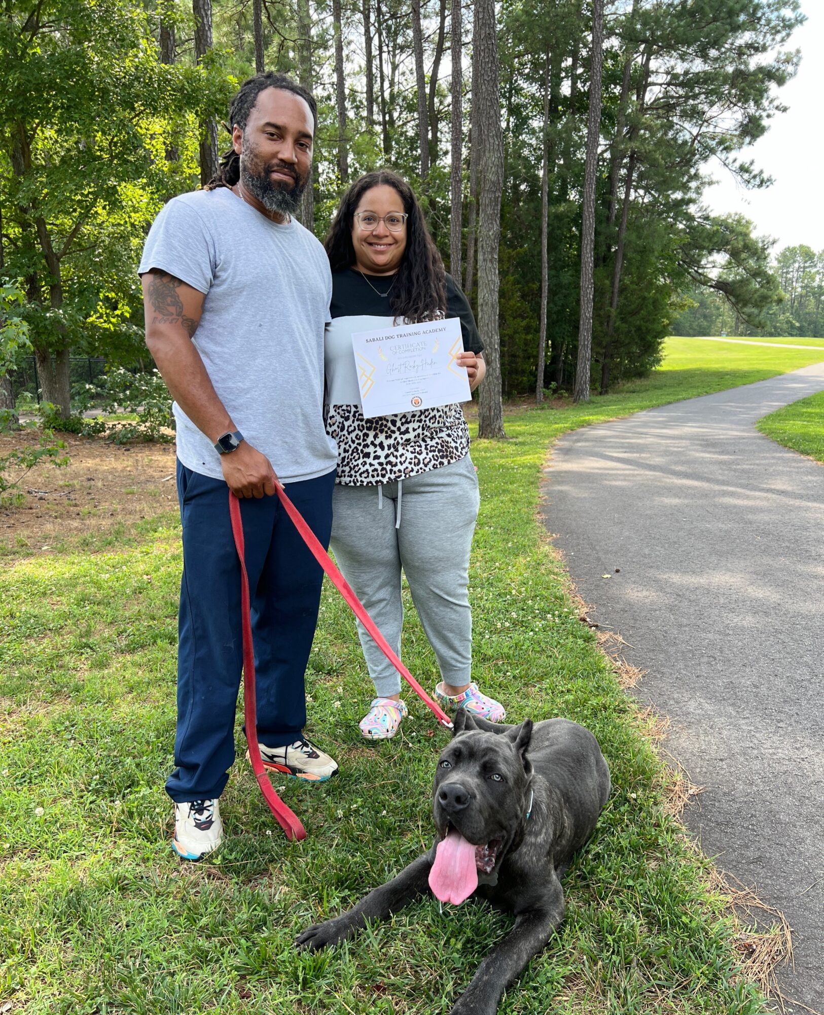 Two people stand beside a gray dog holding a certificate outdoors near a paved path.