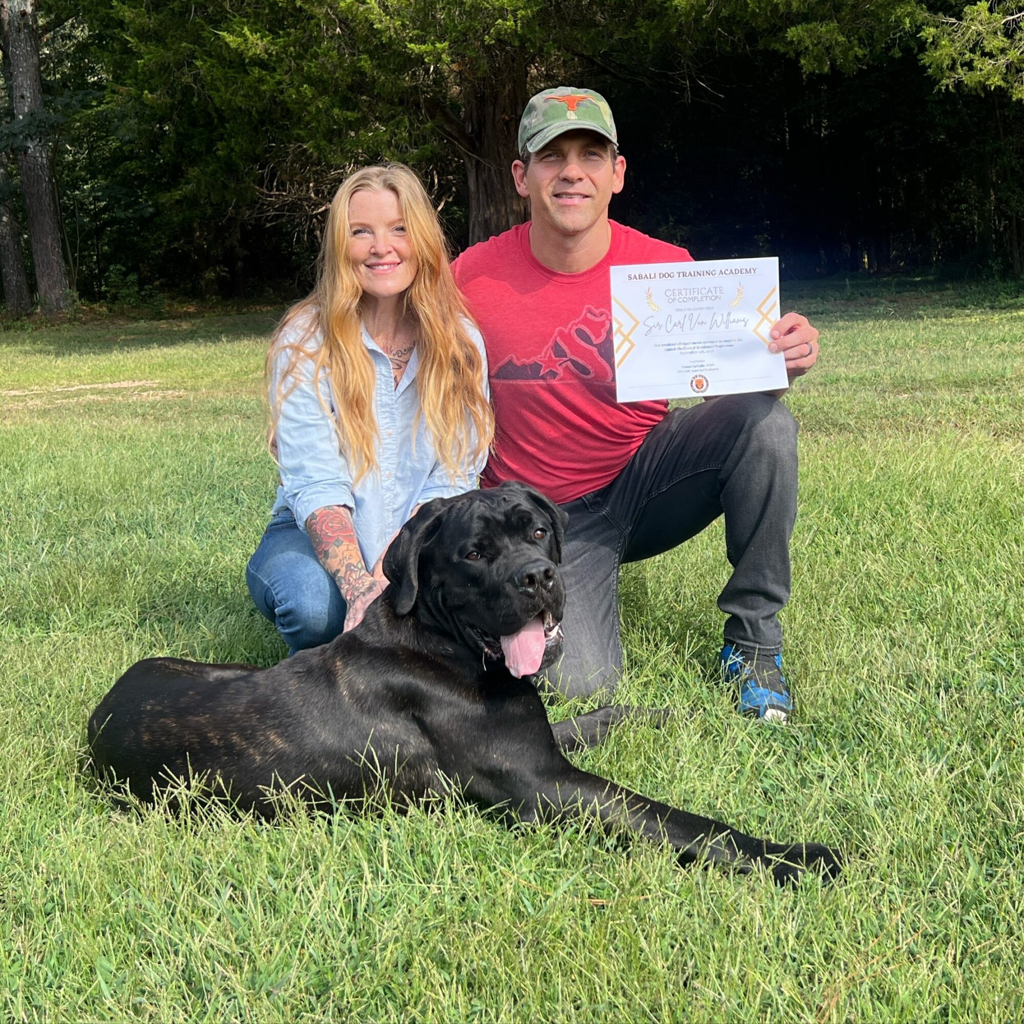 Two people pose outdoors with a large black dog; one holds a certificate.