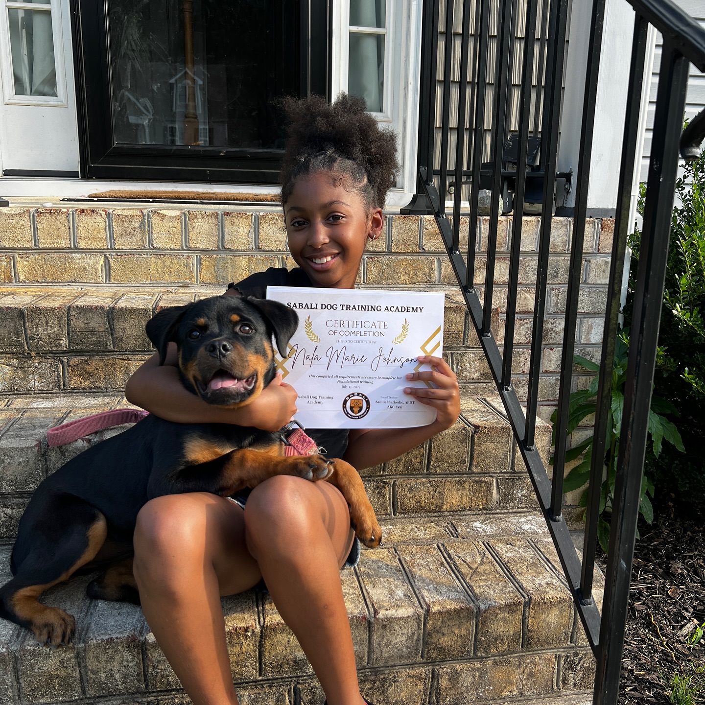 A person seated on stone steps holds a training certificate while embracing a large dog.