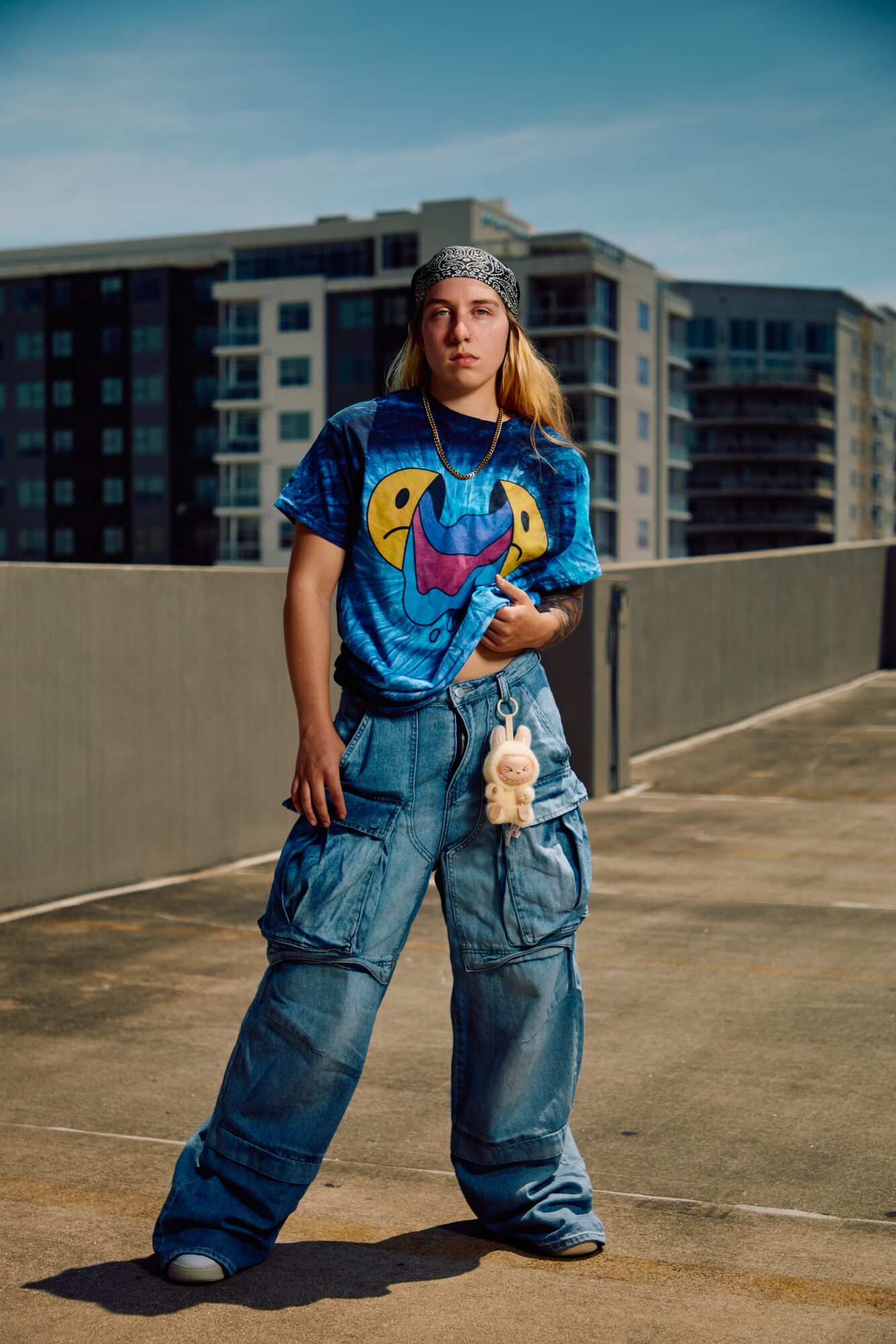 A person wearing a blue graphic shirt, loose jeans, and a patterned head covering stands on a rooftop near apartment buildings.
