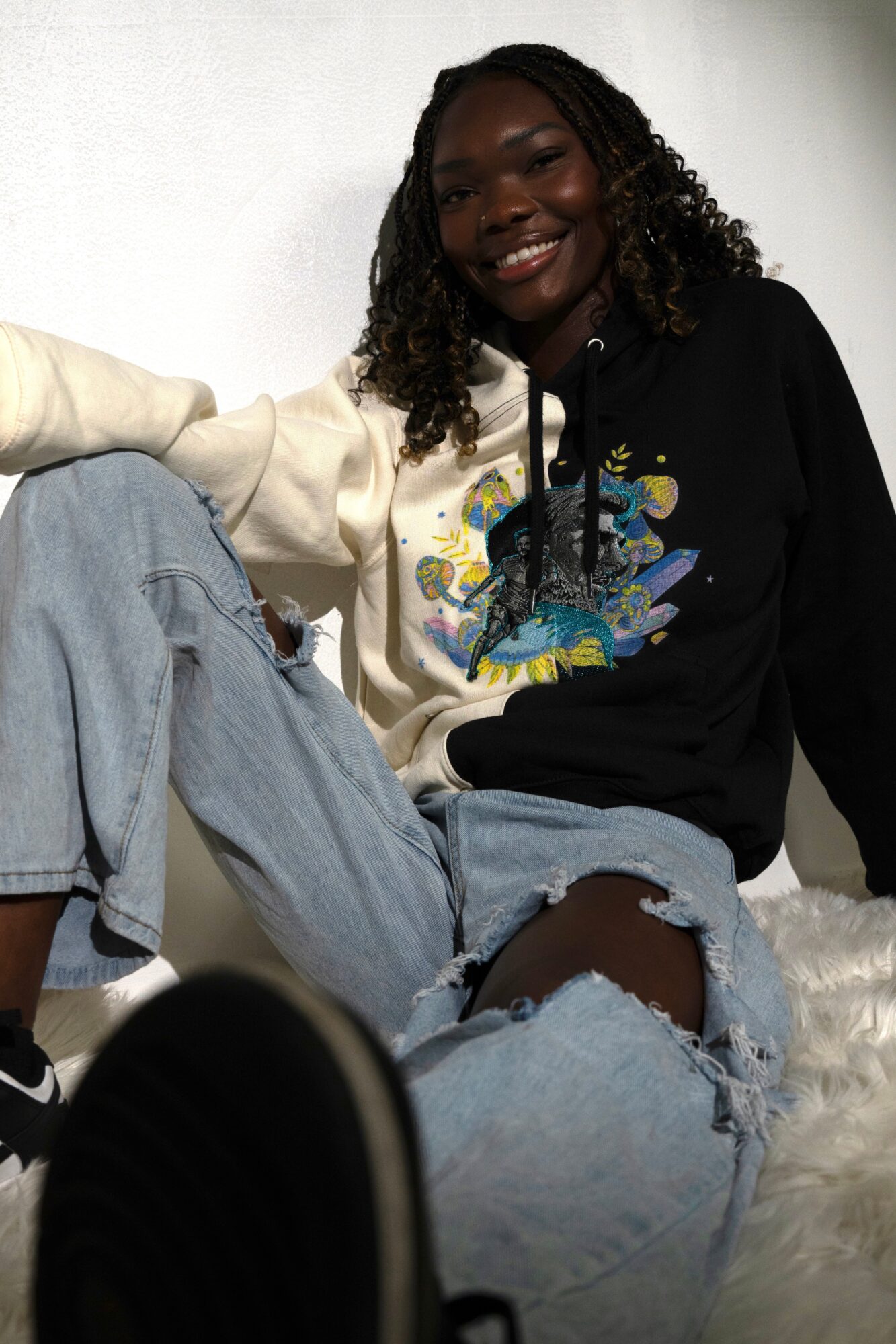 A seated person in jeans and a two-tone graphic hoodie smiles at the camera.