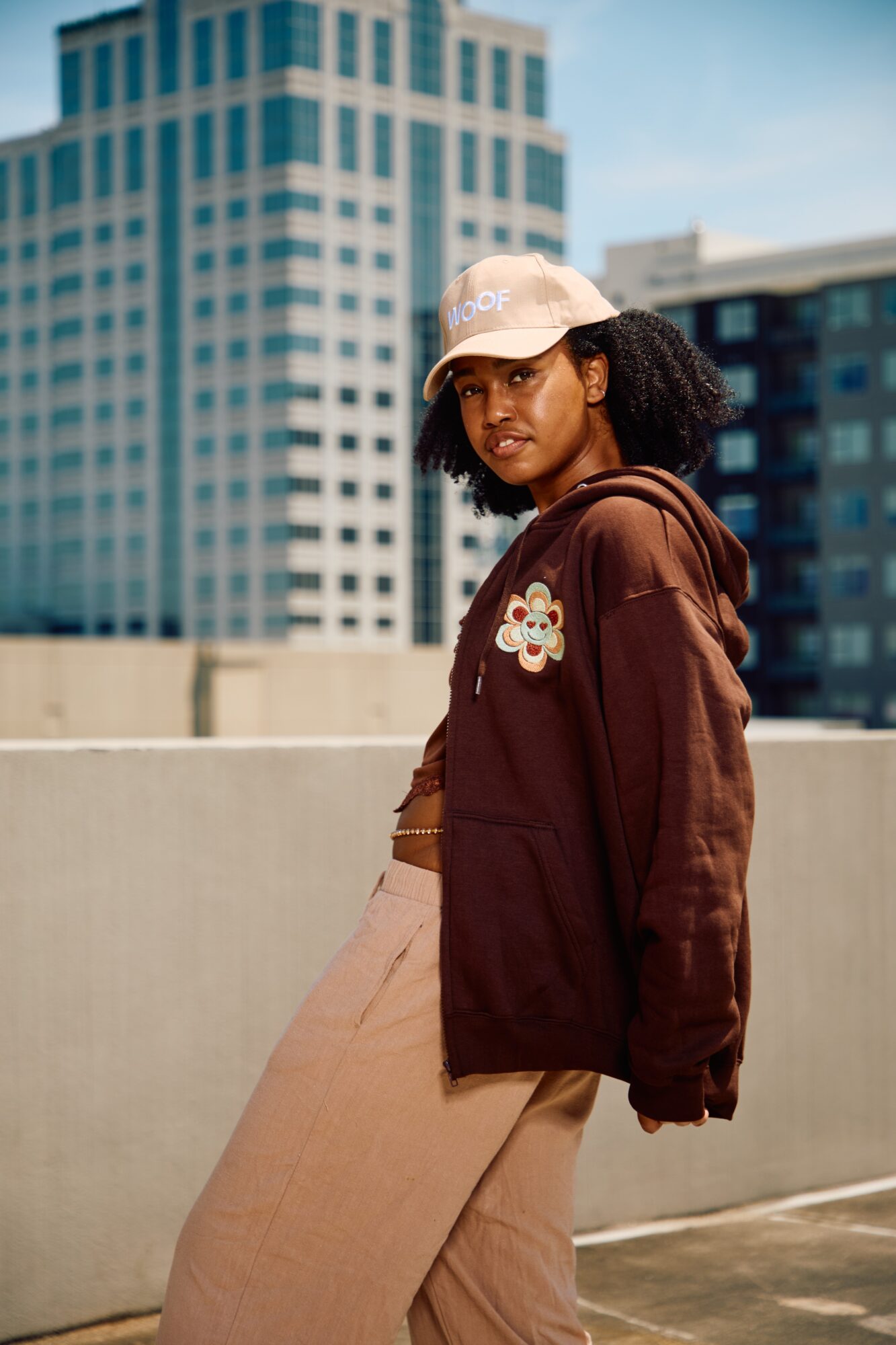 Person in a brown hoodie and light-colored cap standing beside a concrete wall, with tall buildings in the background.