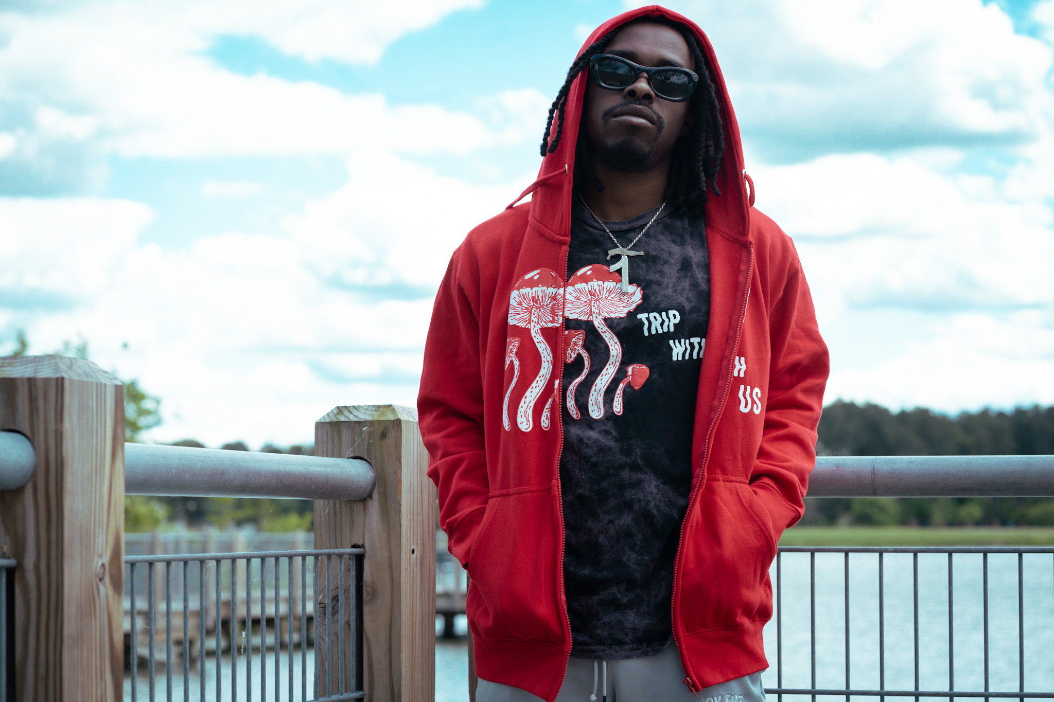 Person wearing sunglasses and a red hoodie stands on a wooden deck beside a metal railing.