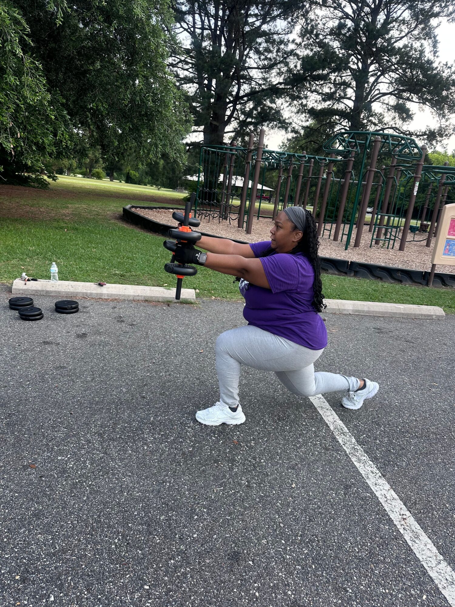 A person in a purple shirt exercises with dumbbells on a paved path beside a grassy area and trees.