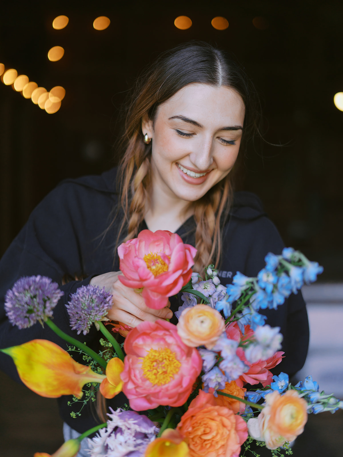 Person smiling while holding a colorful bouquet of flowers against a dark background with warm lights.