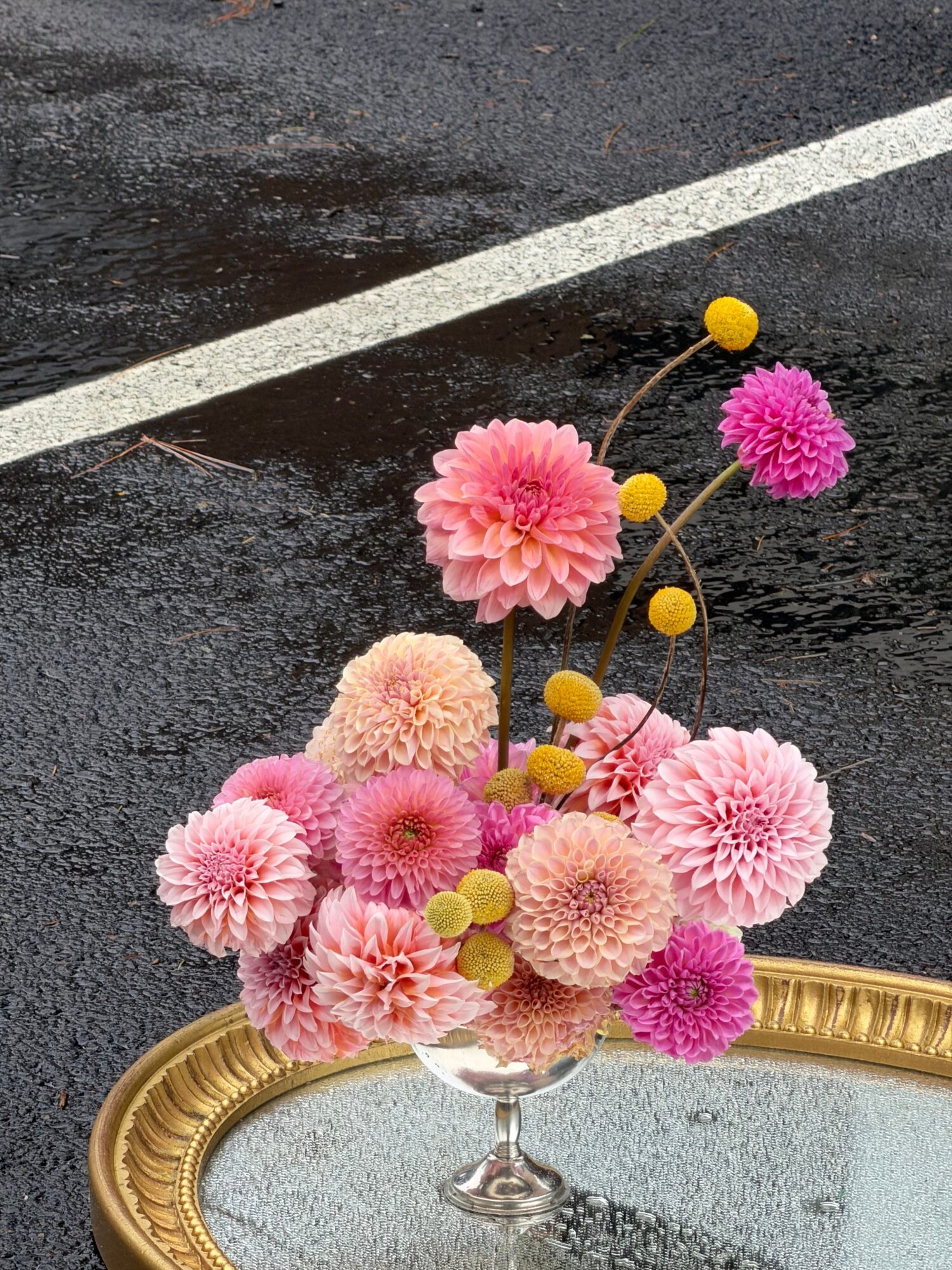 Pink flowers and yellow berry-like accents arranged in a glass vase beside an ornate mirror.