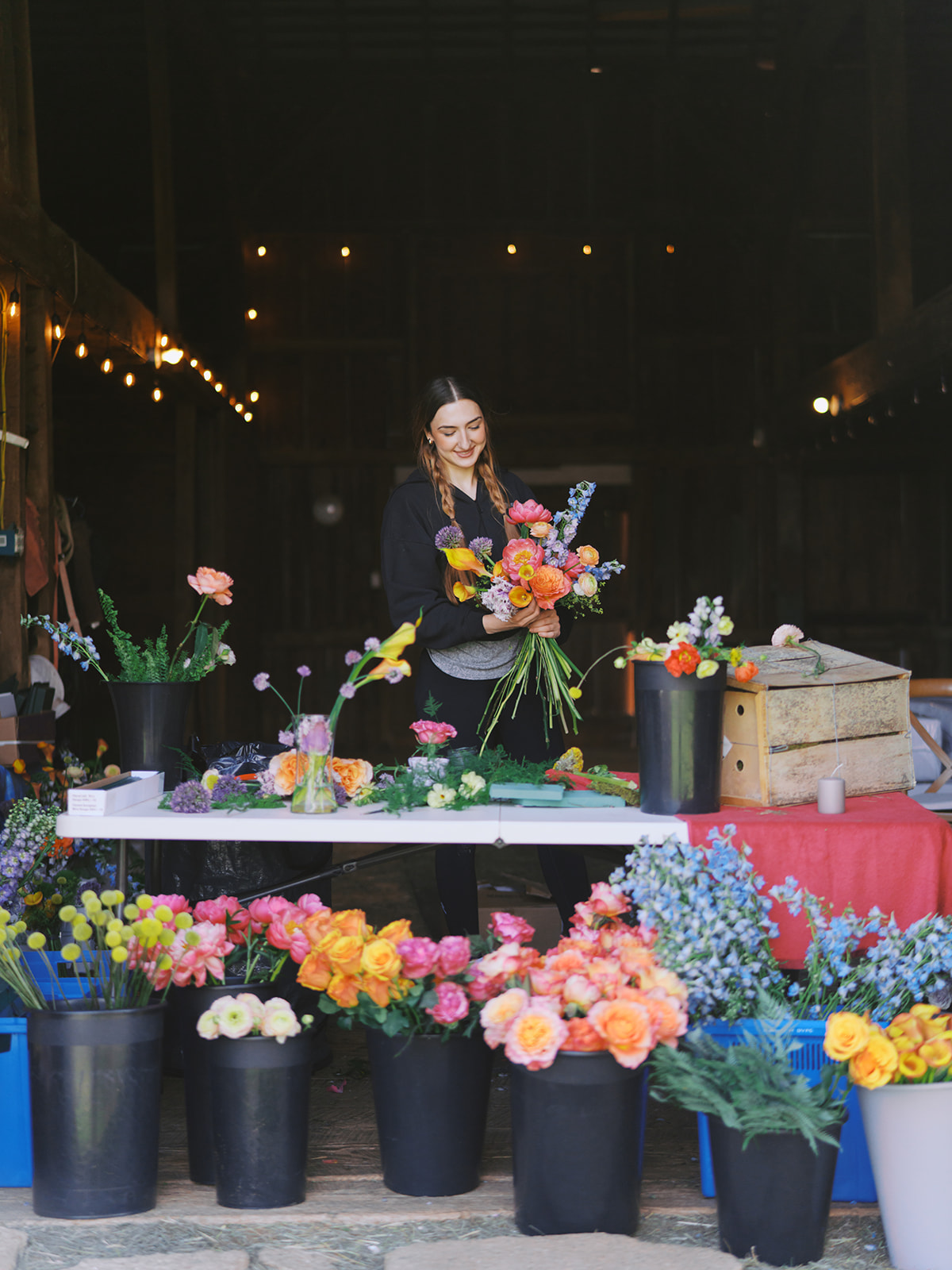 A person arranges a colorful bouquet behind a flower stall filled with buckets of flowers.