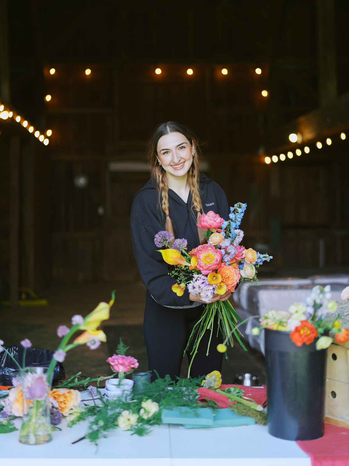 Person holding a colorful bouquet behind a flower-covered table in a dimly lit indoor space.