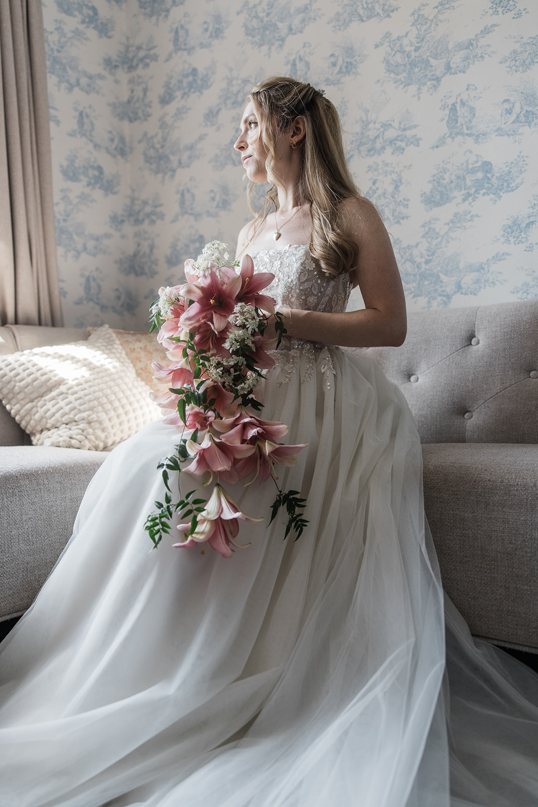 A person in a white gown holds a cascading bouquet beside a gray sofa.