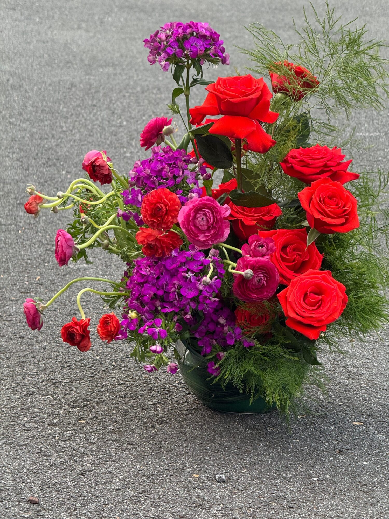 A colorful bouquet of red roses and purple flowers rests on a gray pavement surface.