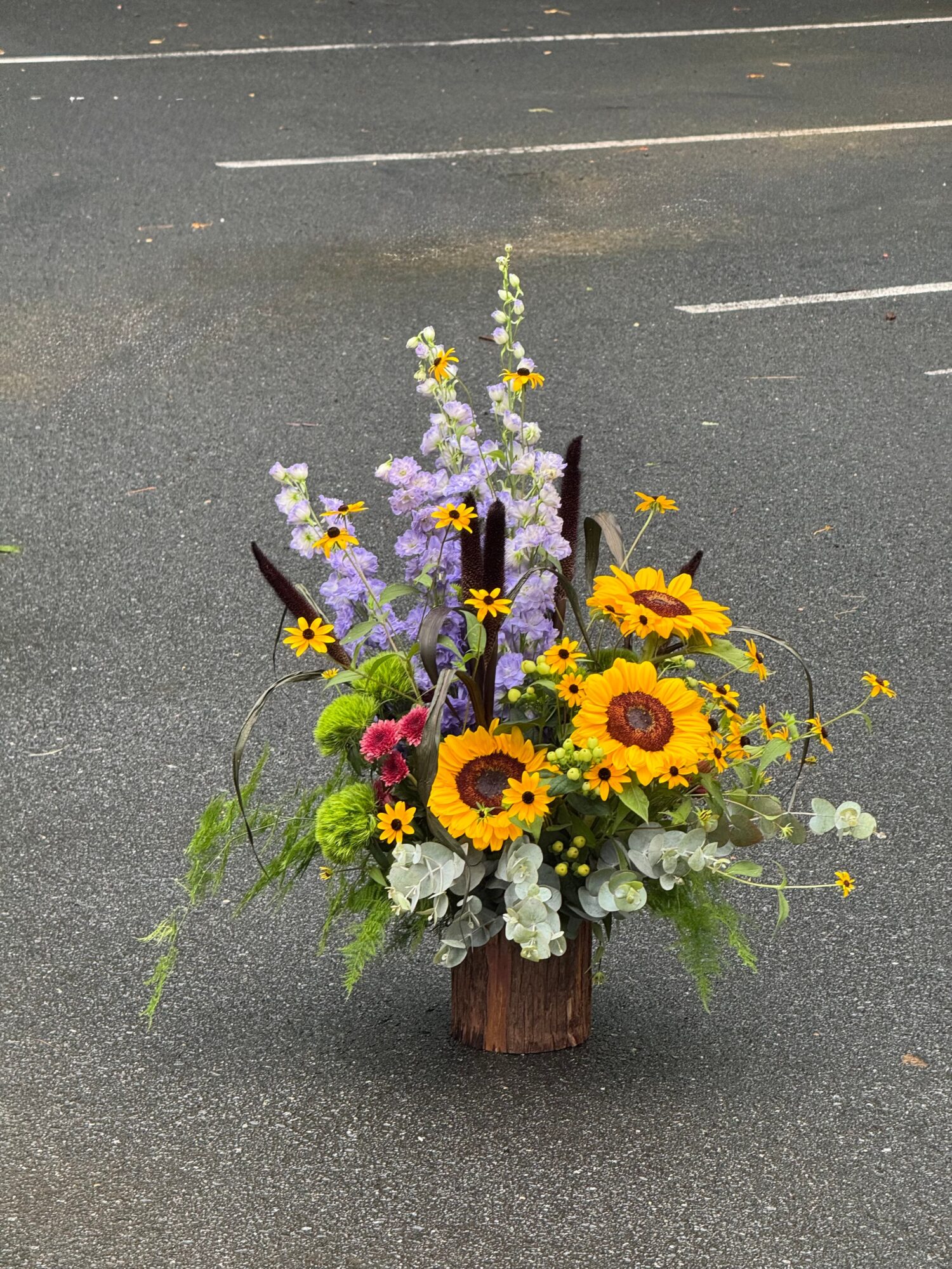 A colorful arrangement of yellow, purple, and pink flowers in a wooden container on asphalt.