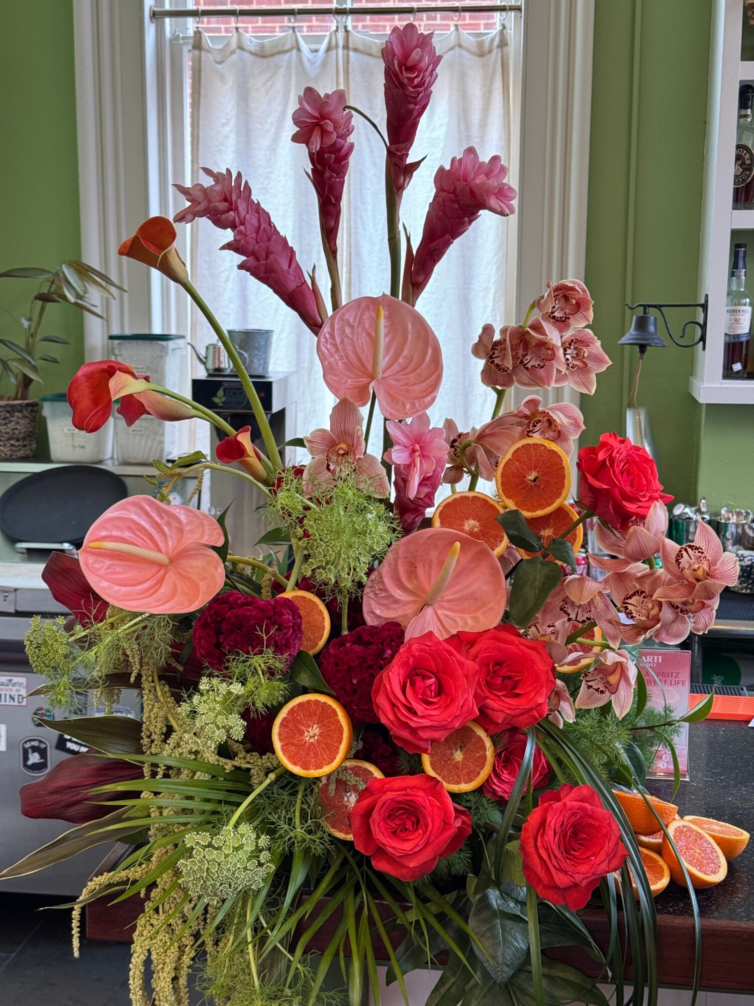 A colorful floral arrangement with red roses, pink anthuriums, orange accents, and greenery beside a window.