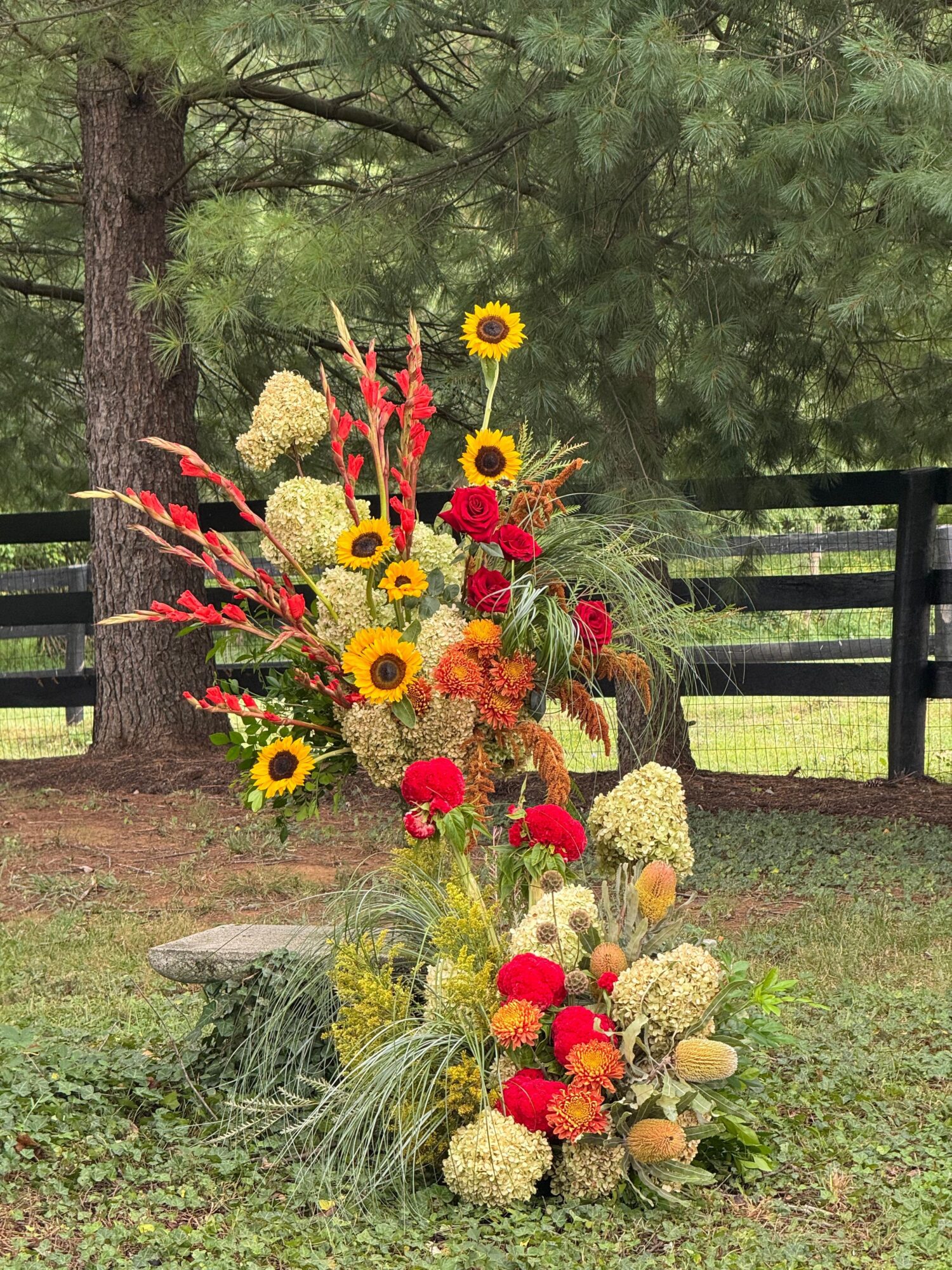 Colorful flower arrangements rest on a wooden bench beside a black metal fence and evergreen branches.