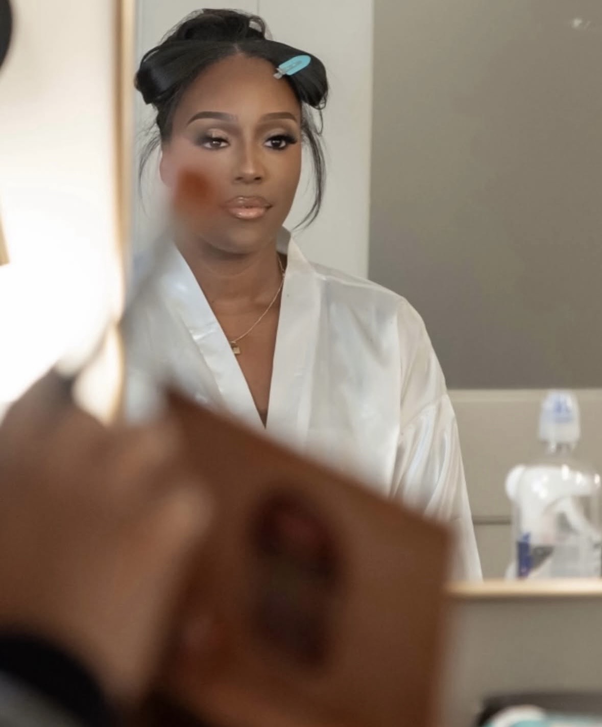 A person in a white robe looks into a mirror while applying makeup.