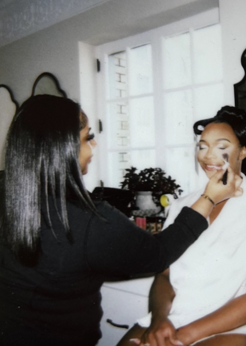 A person applies makeup to another person seated in a white robe near a bright window.