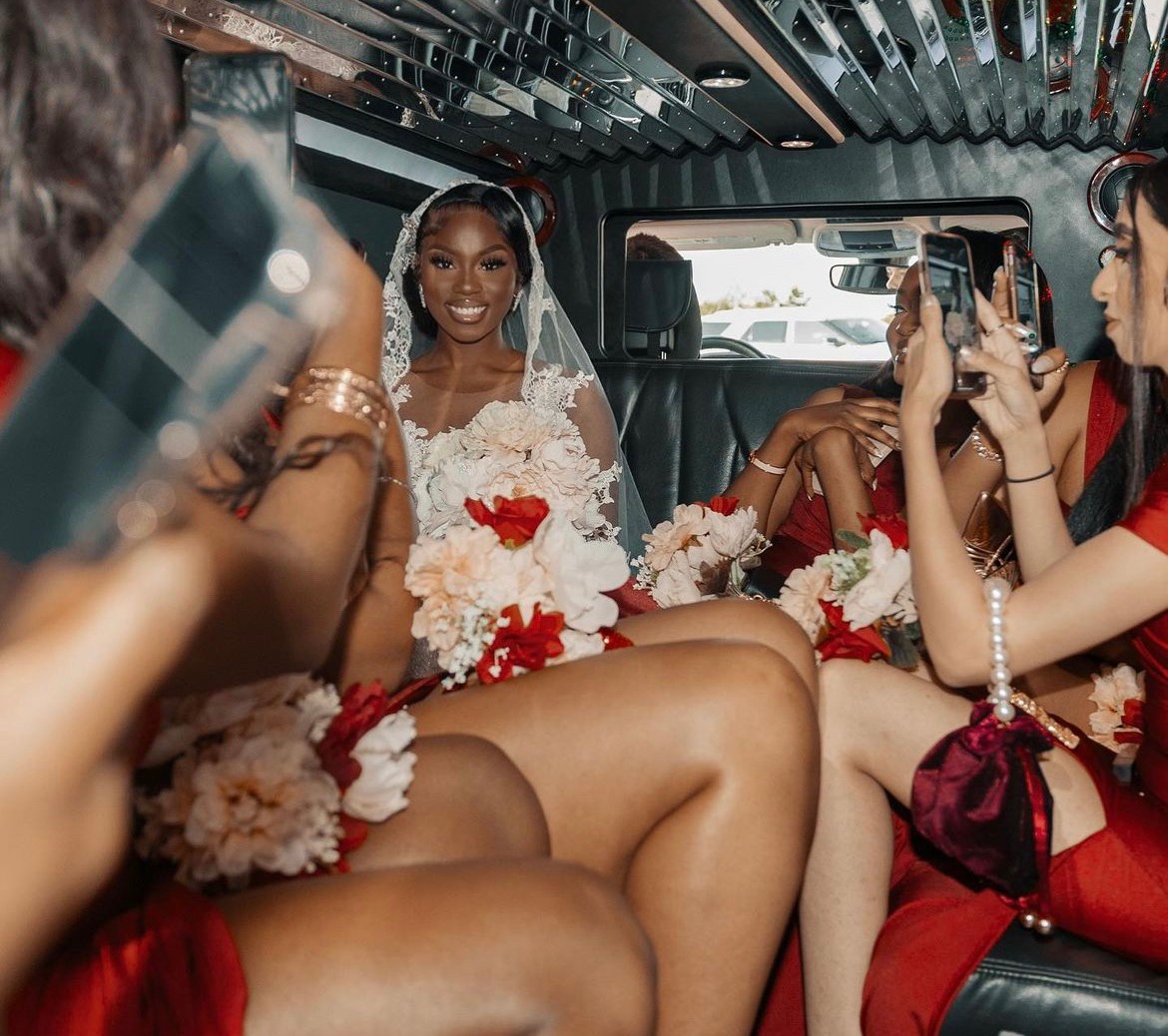 A bride in a white dress sits in a limousine among people holding phones and bouquets.