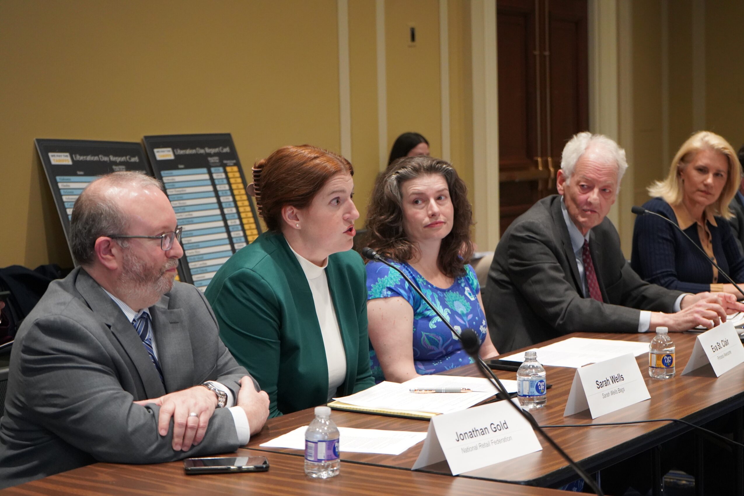 Five people sit at a conference table with papers, microphones, and water bottles; presentation boards stand behind them.