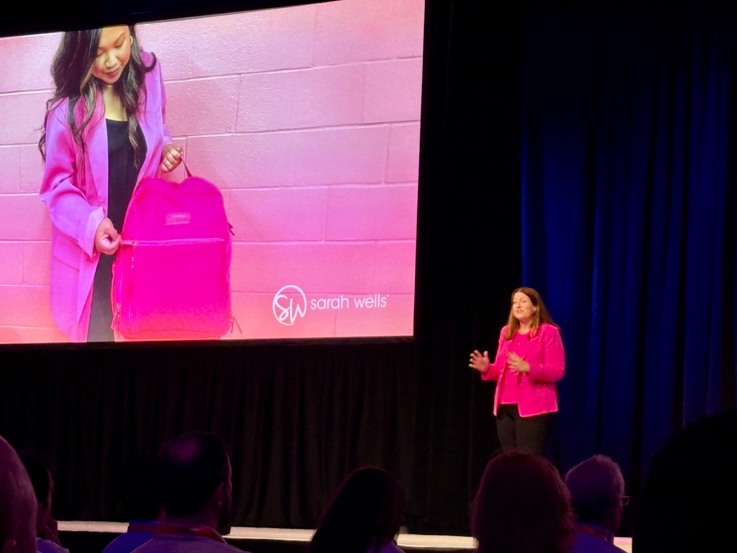 A presenter in a bright pink jacket stands beside a projected image of a person with a pink suitcase, facing an audience.