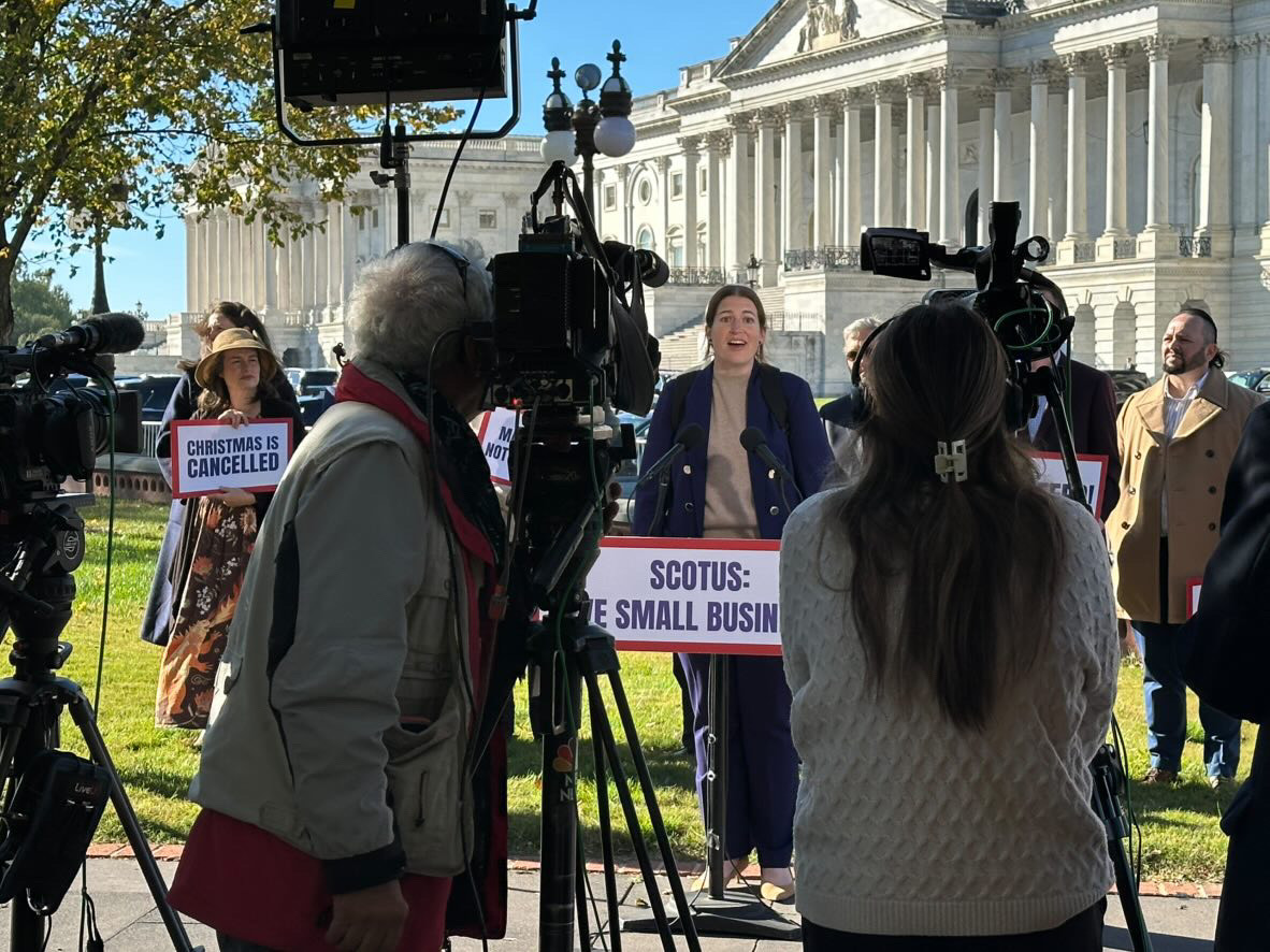 People and television cameras gather around a woman speaking outdoors in front of a columned building.