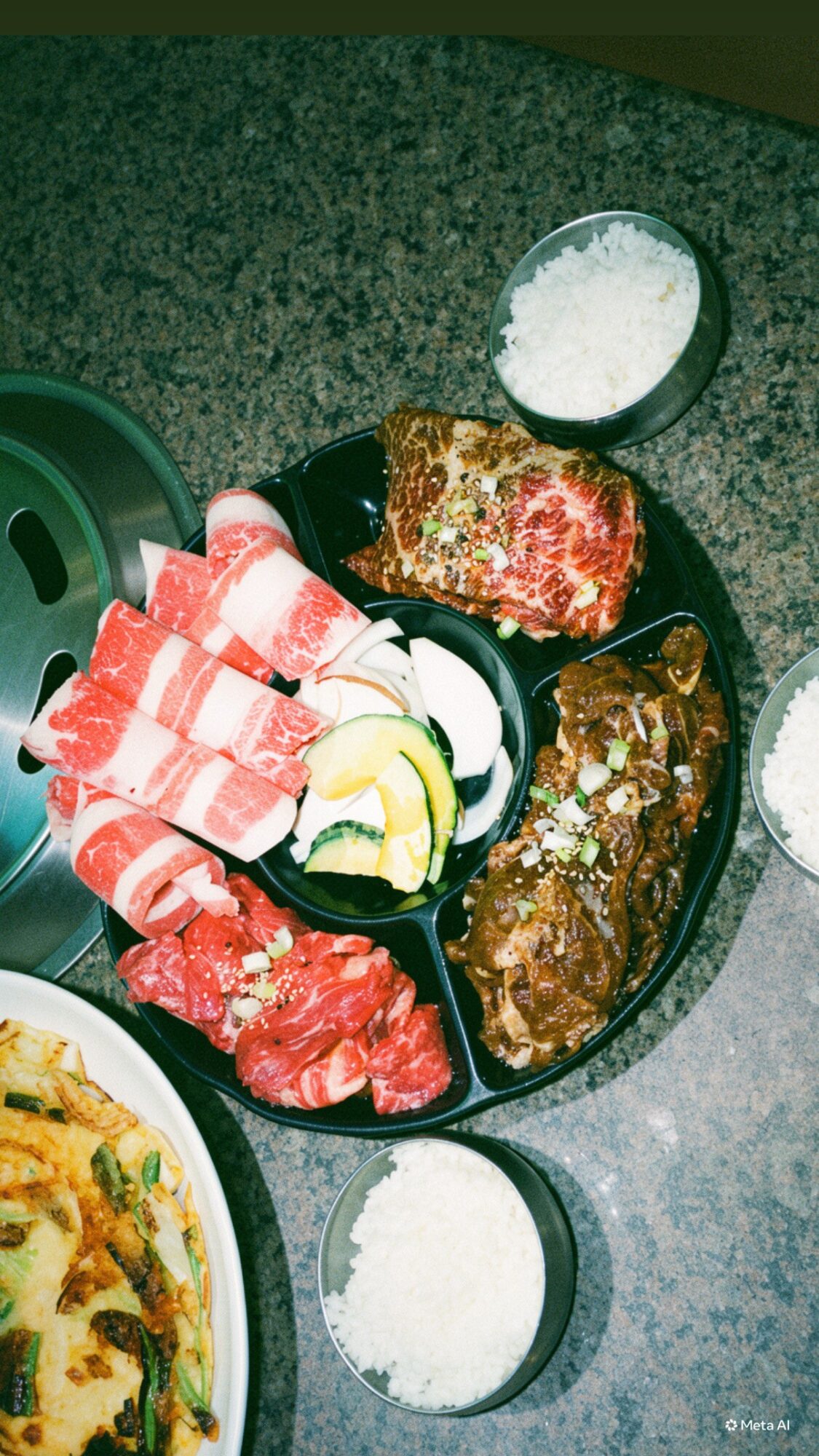 A divided serving platter with sliced meat, salad, and cooked dishes, surrounded by bowls of rice on a countertop.