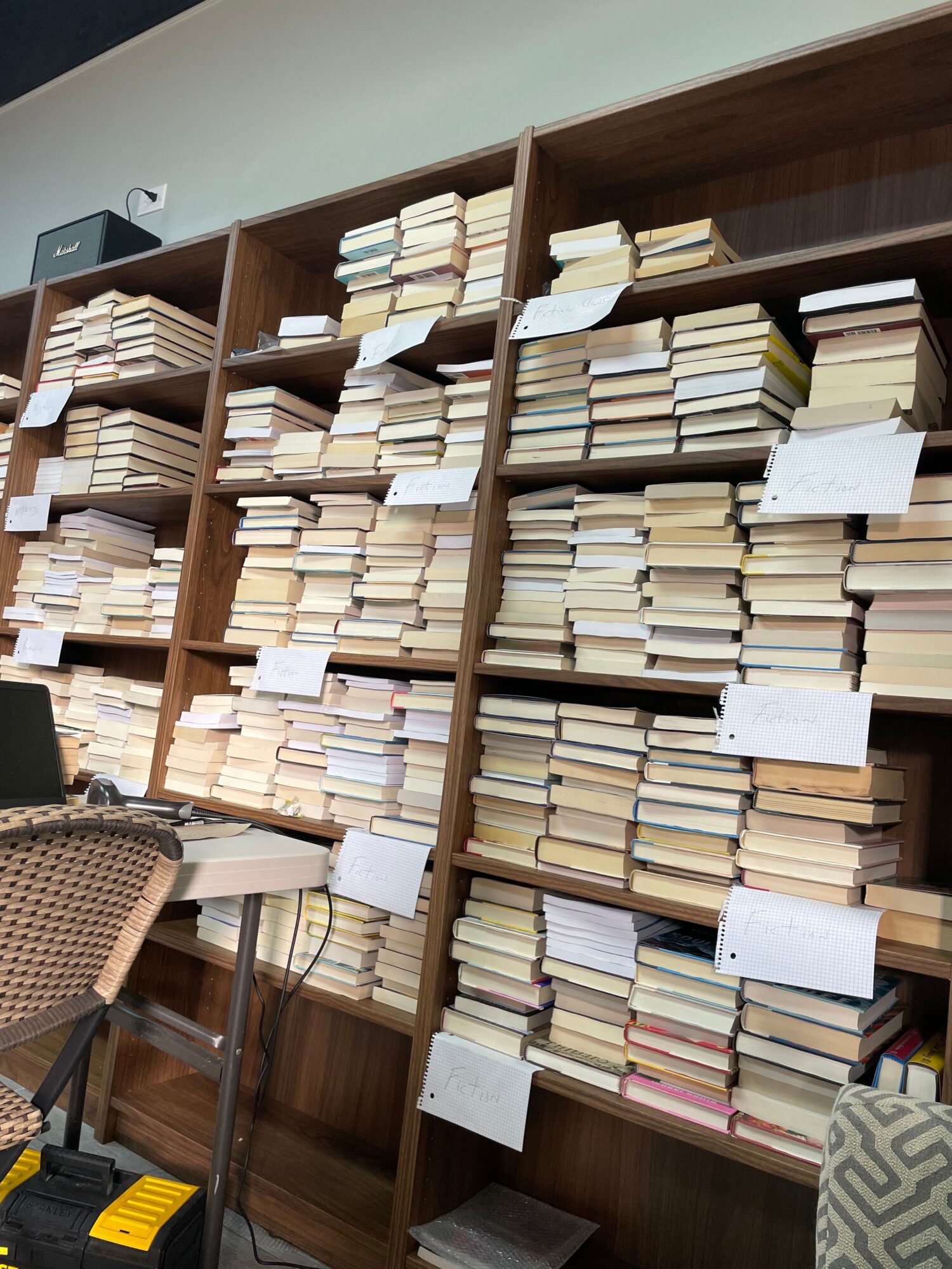 Tall wooden shelving filled with books and papers, with a woven chair and a yellow object at the lower right.