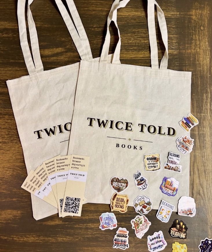 Two canvas tote bags labeled “TWICE TOLD BOOKS” with bookmarks and colorful stickers on a wooden table.