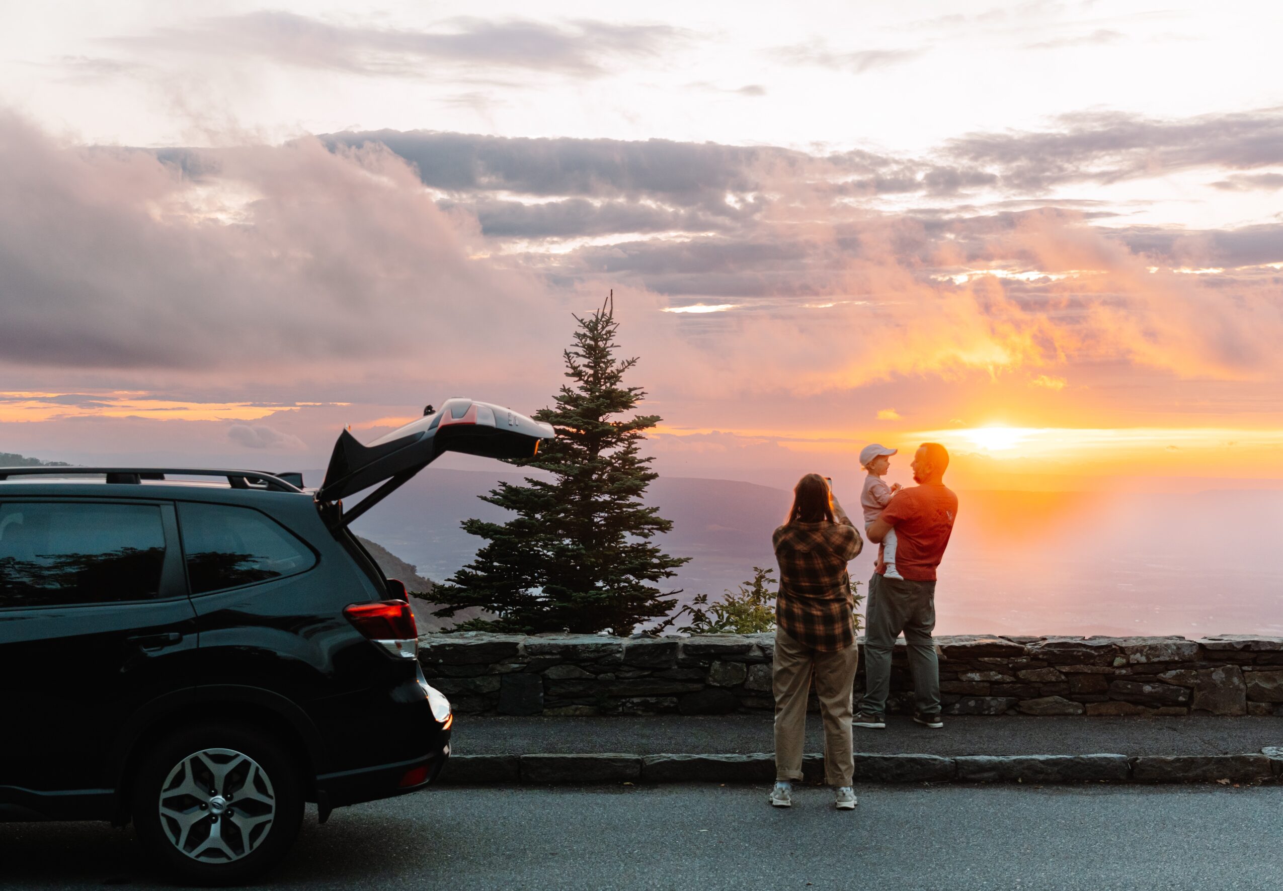 People stand beside a parked car and stone wall, watching a sunset over the water.