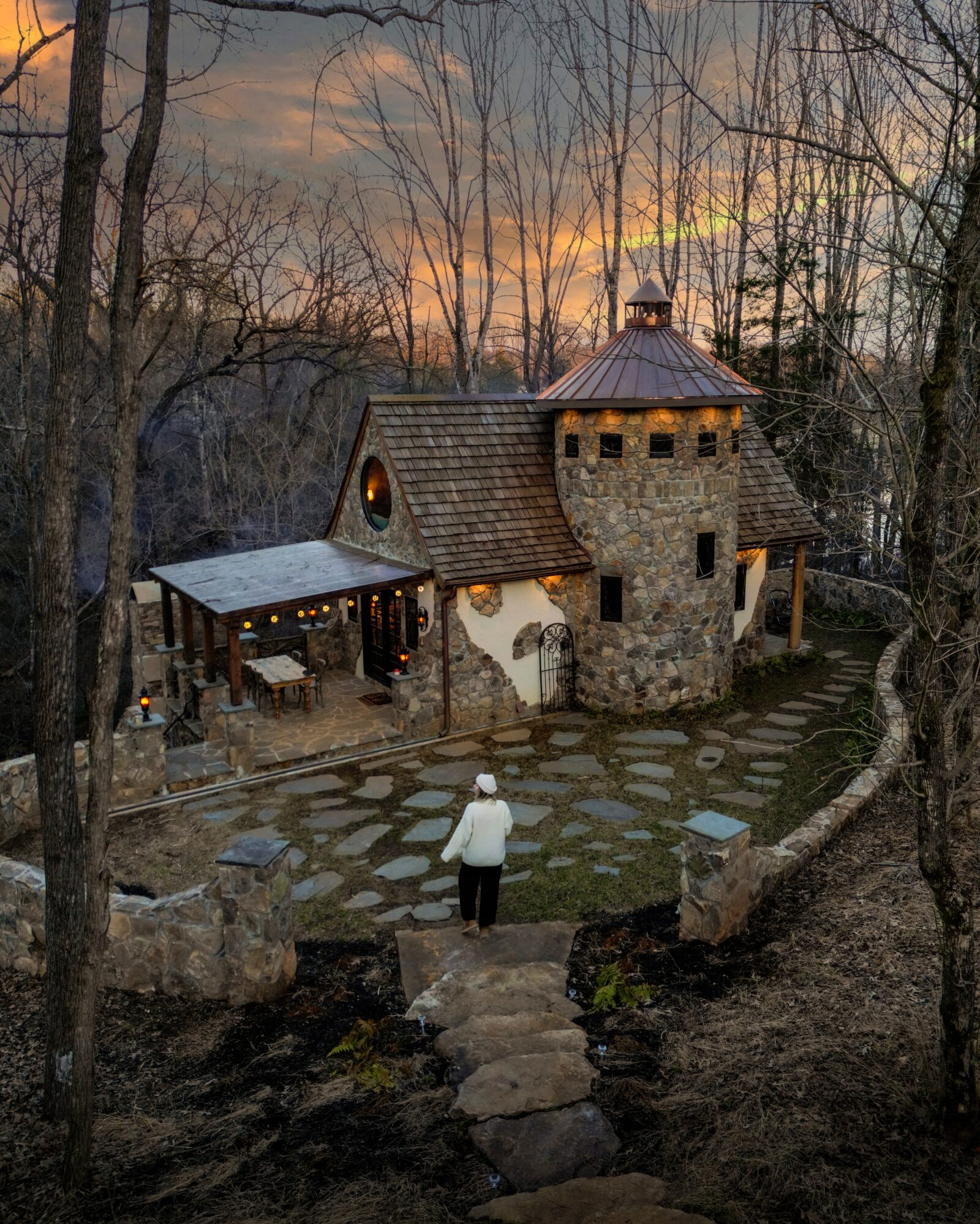 Stone building among leafless trees at sunset, with a person standing on a stone path in the foreground.