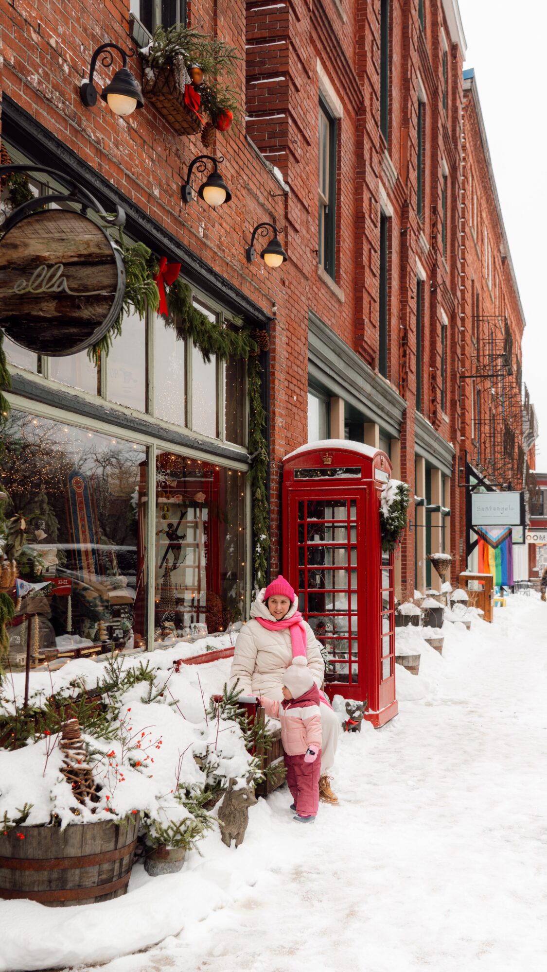 Person in winter clothing walks through snow beside a red telephone booth and brick storefronts.