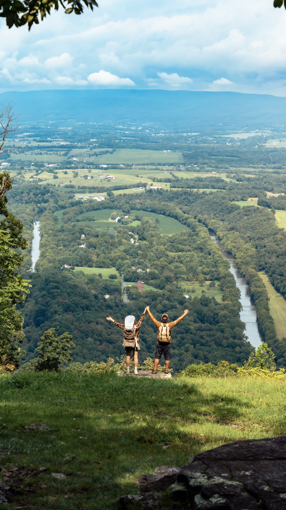 Two people stand at a grassy overlook above a forested valley and winding river.