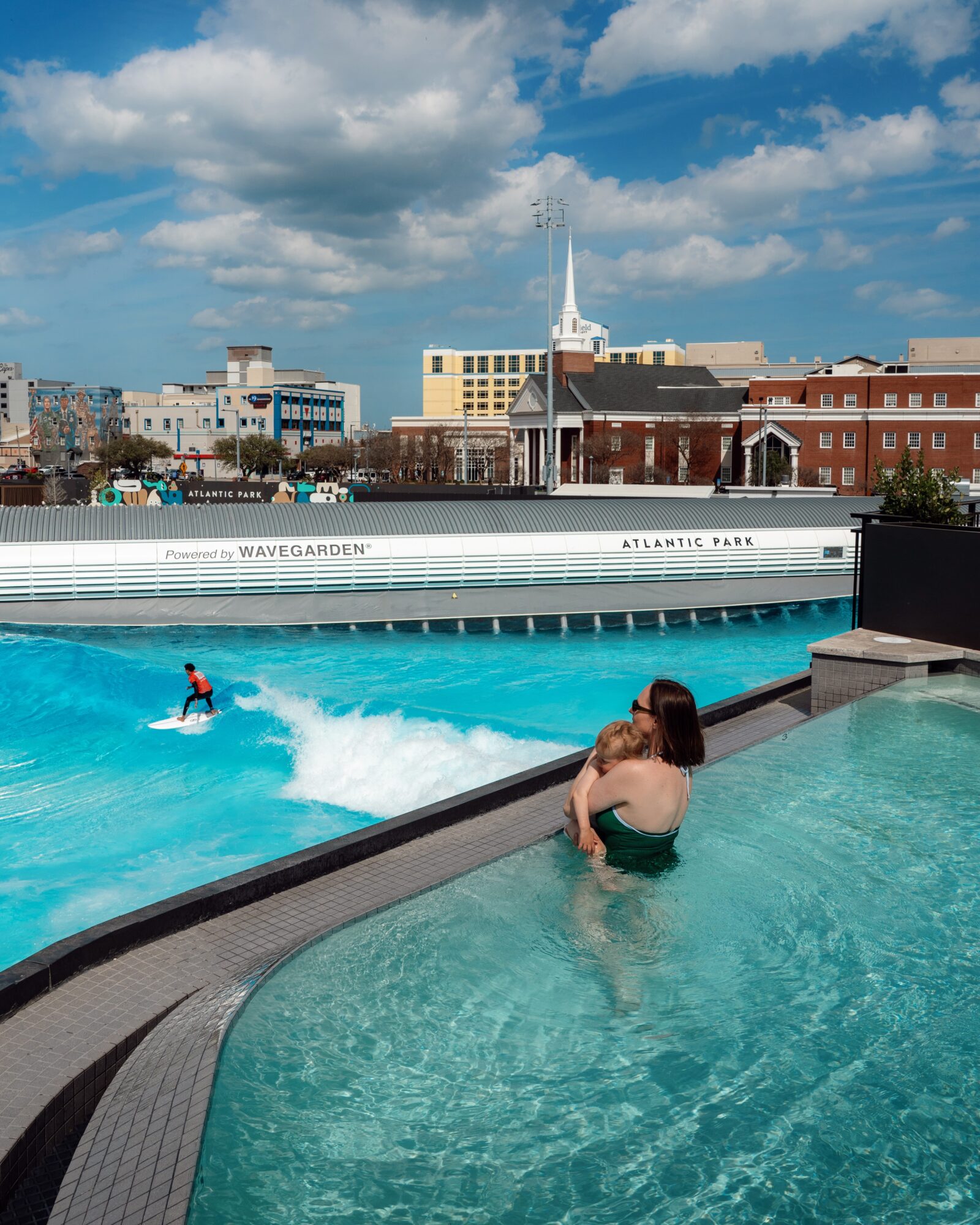 A person sits in a shallow pool beside a larger wave pool, with city buildings and a cloudy sky behind them.