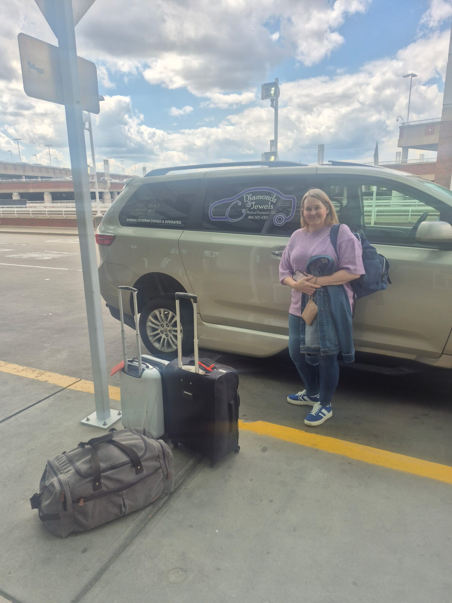A person stands beside a parked minivan, with several bags on the pavement nearby.