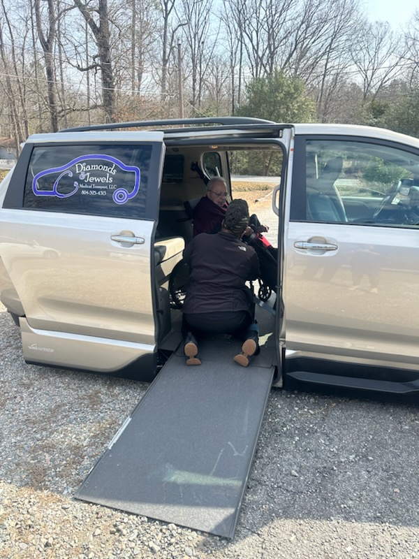 An open minivan with a ramp extending to the ground as two people assist someone inside.