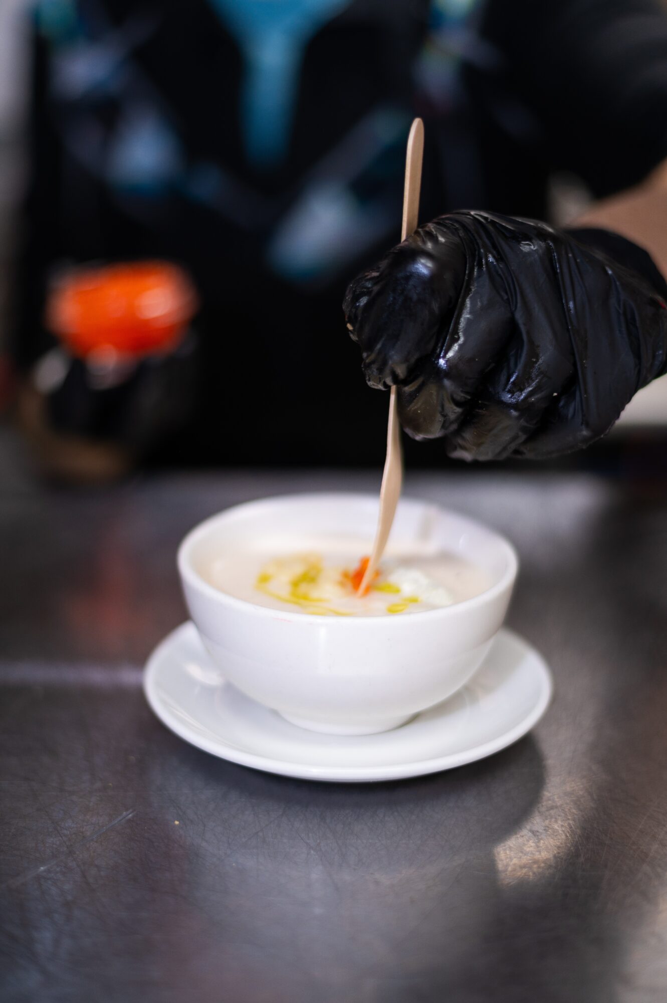 A gloved hand stirs a small white bowl of food with a wooden stick on a countertop.