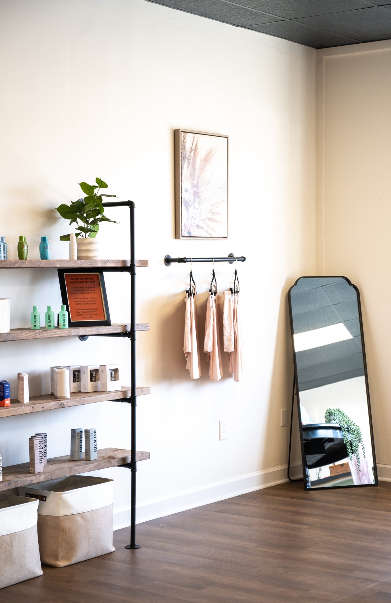 A bright room with wall shelves, potted plants, framed artwork, hanging garments, and a leaning full-length mirror.
