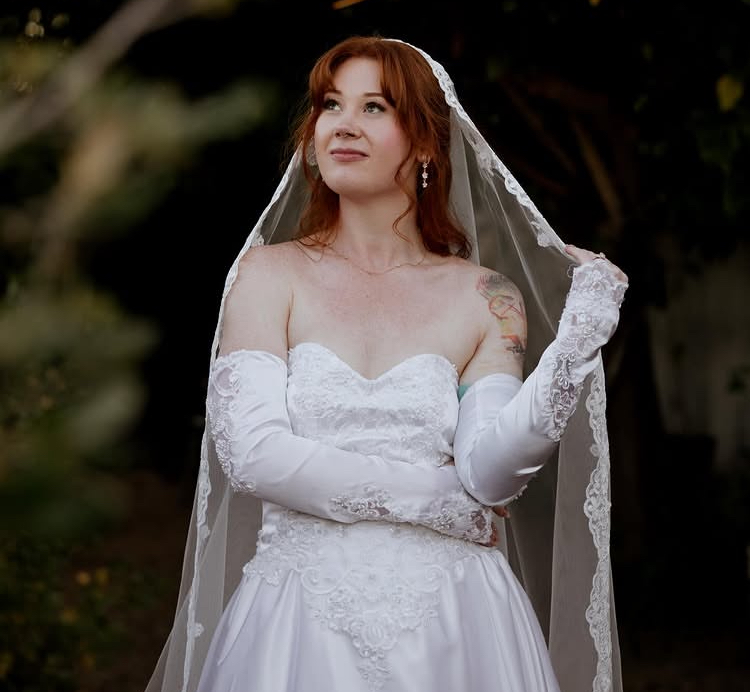 A red-haired person wearing a white wedding dress, veil, and long gloves outdoors.