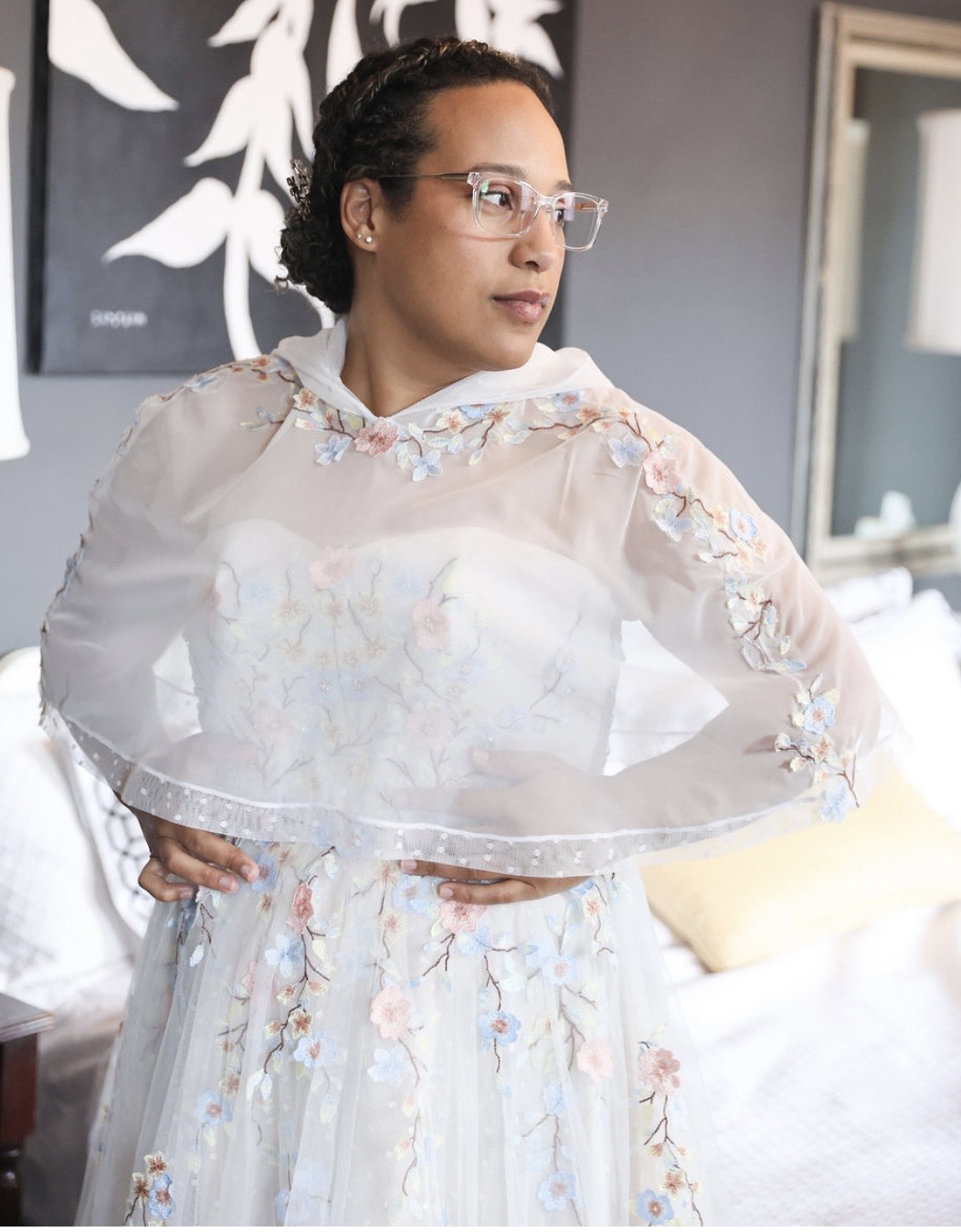 Person wearing glasses and a pale embroidered outfit stands beside a bed, looking to the side.