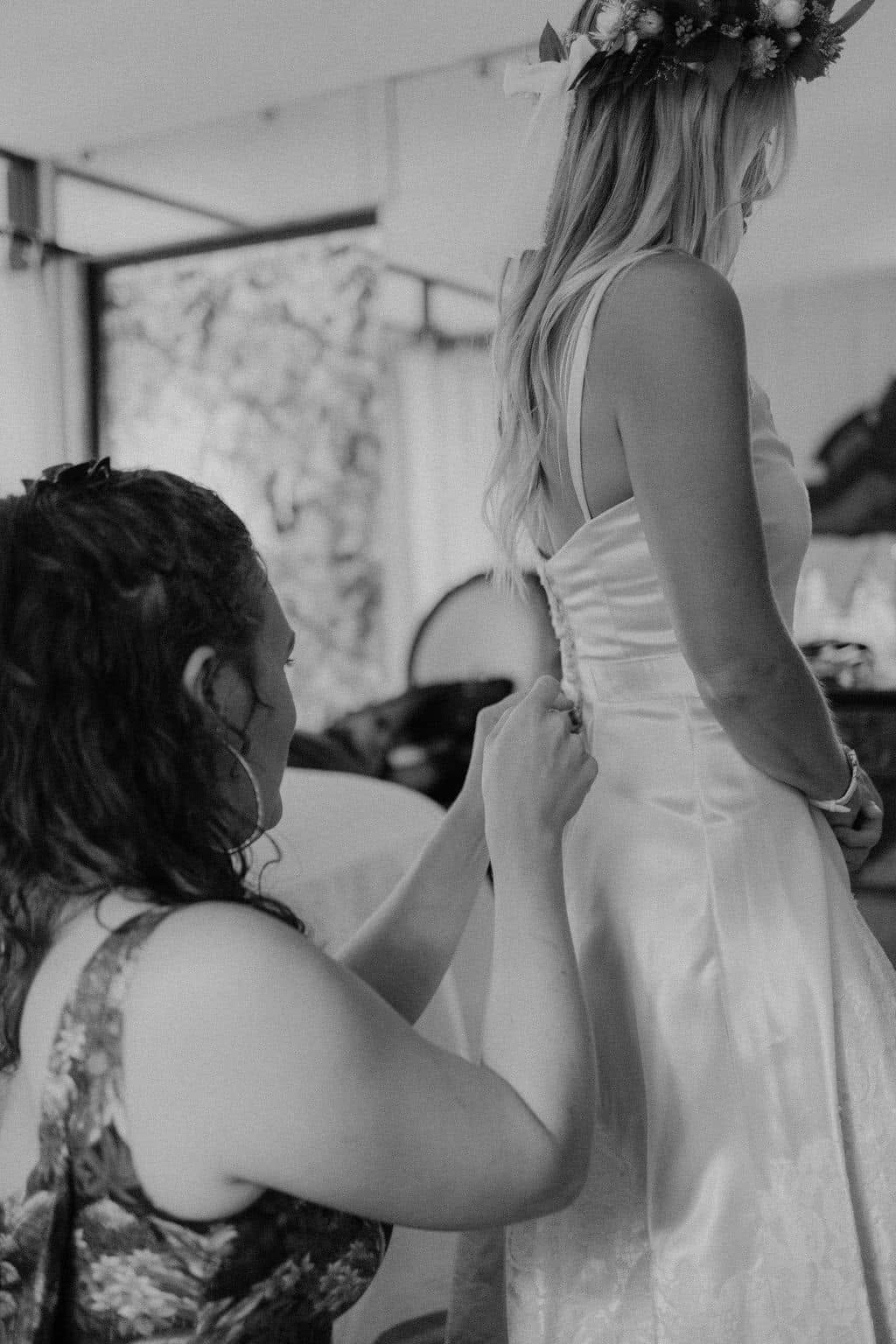 A seated person adjusts the back of a standing person's white dress beneath a floral headpiece.