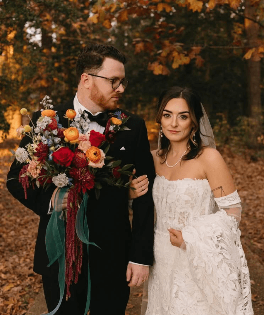 A person in a dark suit holding a colorful bouquet stands beside a person in a white wedding dress outdoors.