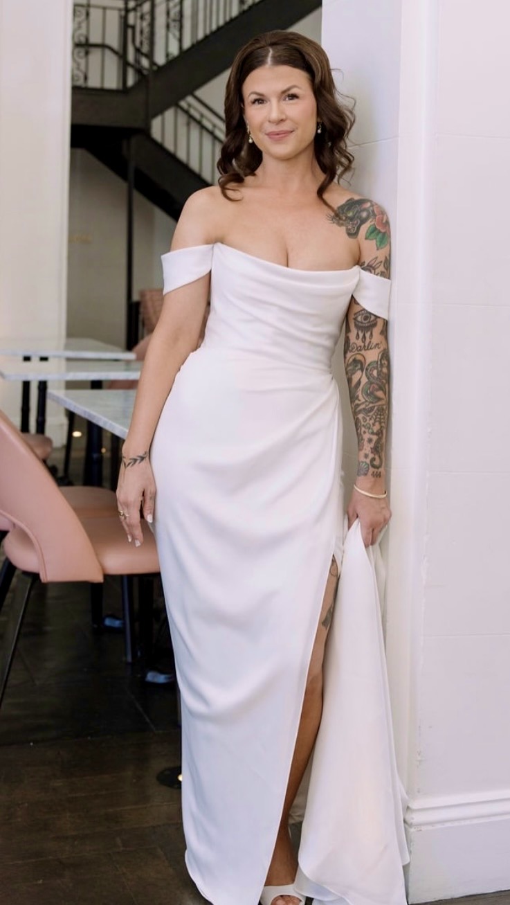 Person in a white gown standing indoors beside a staircase.