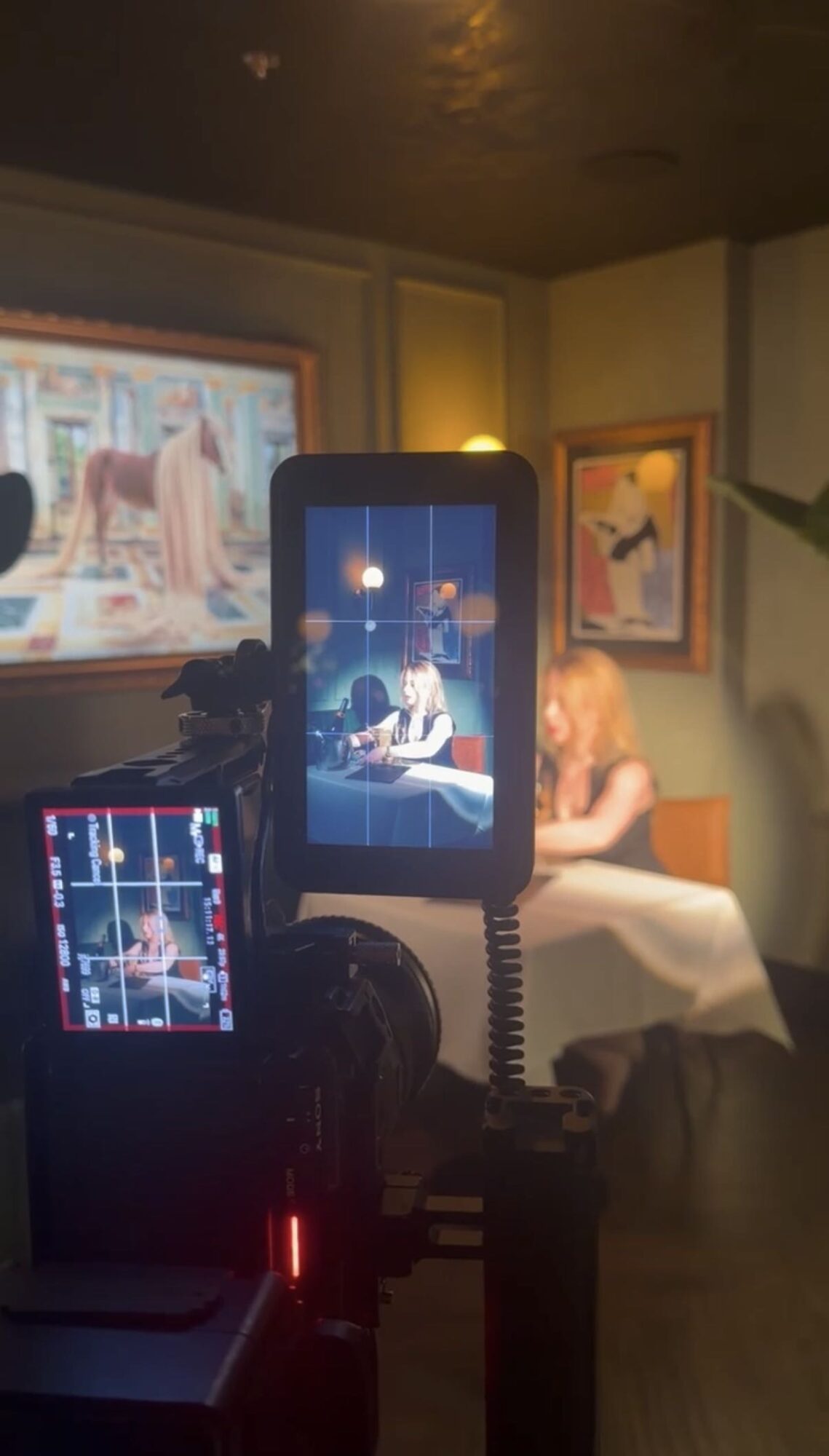 A camera setup films a seated person at a table in a room with framed artwork.