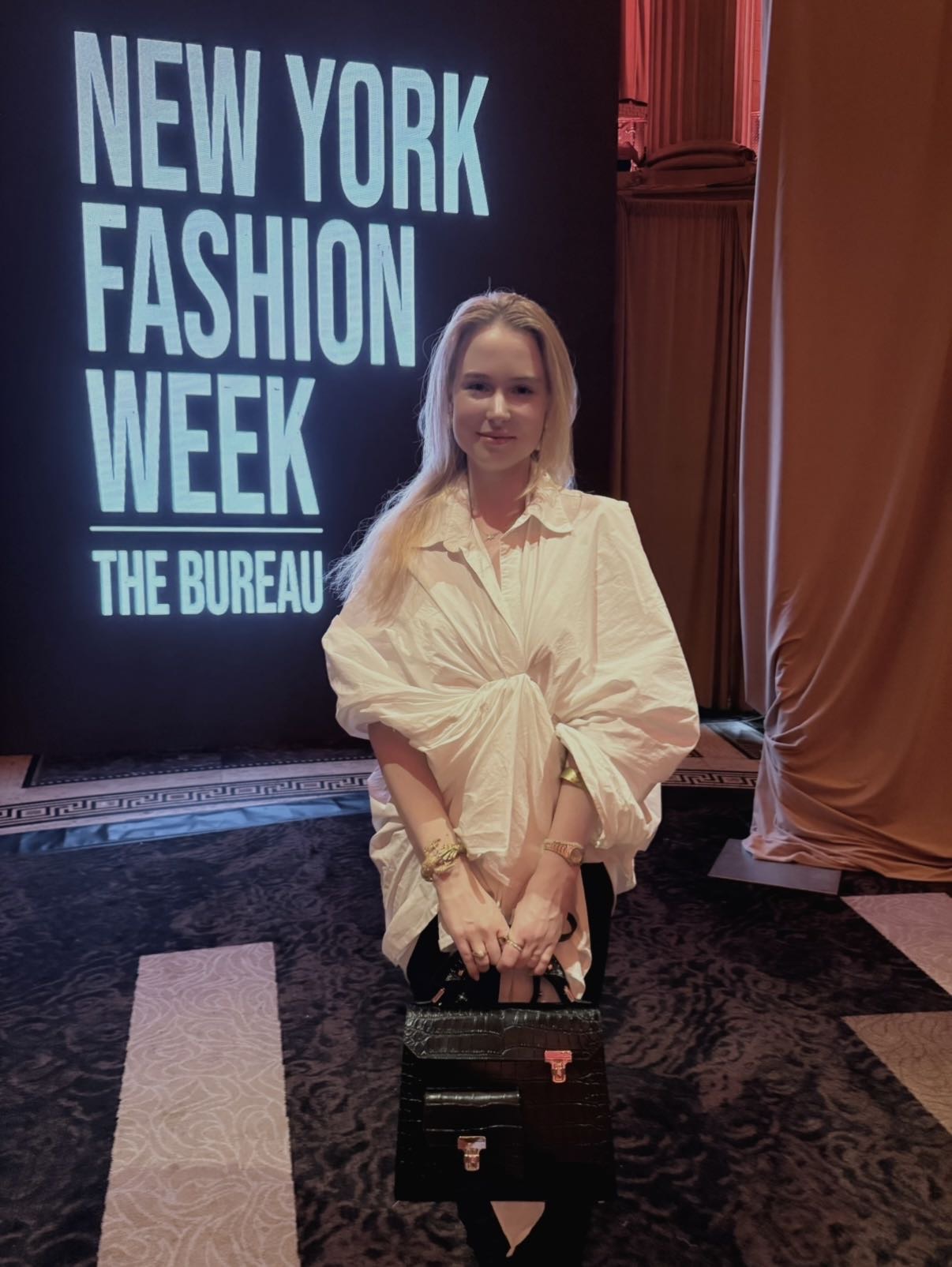 A person stands in front of a New York Fashion Week sign, holding a black handbag.