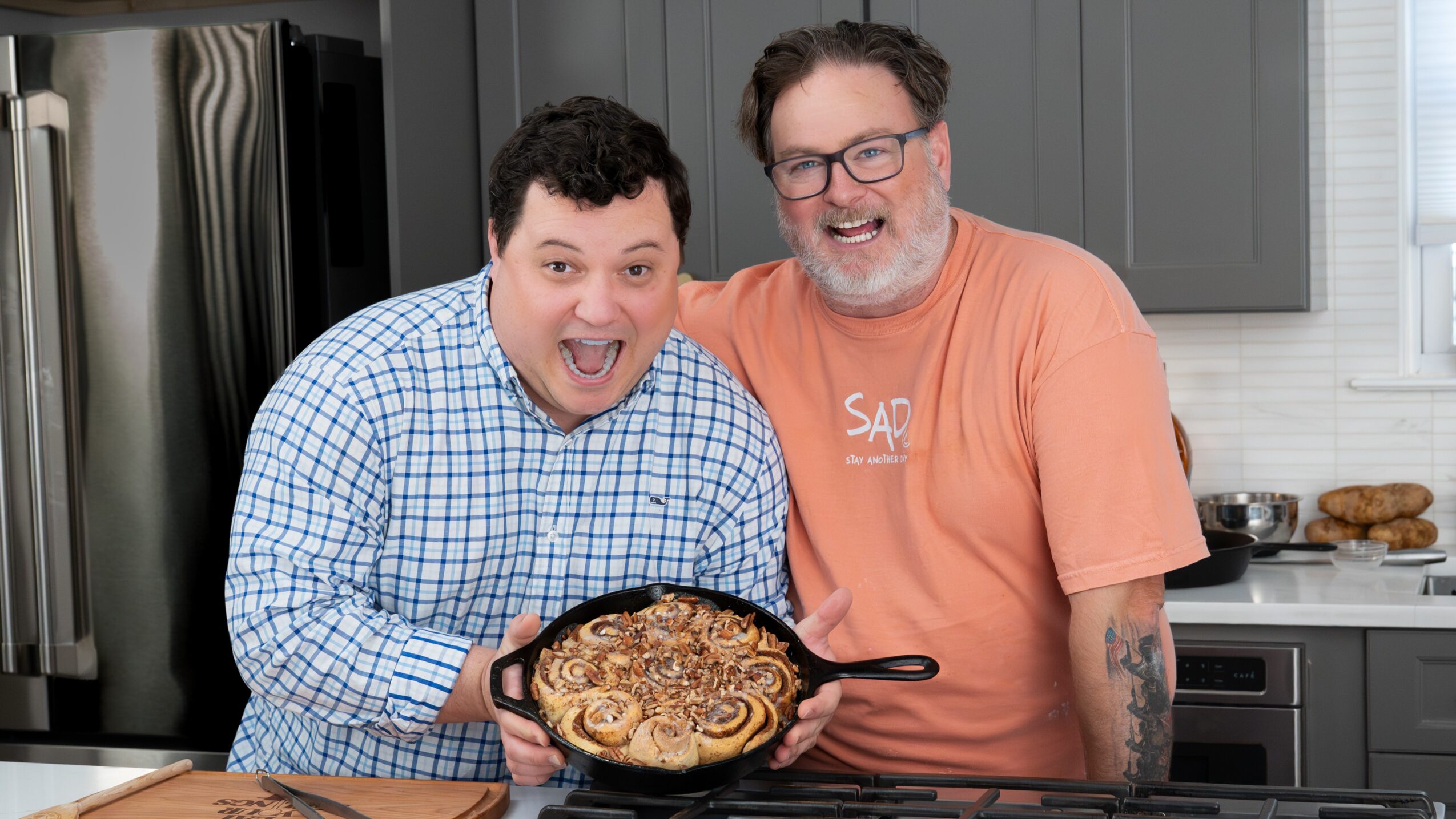 Two people stand in a kitchen, holding a pan of cooked food.