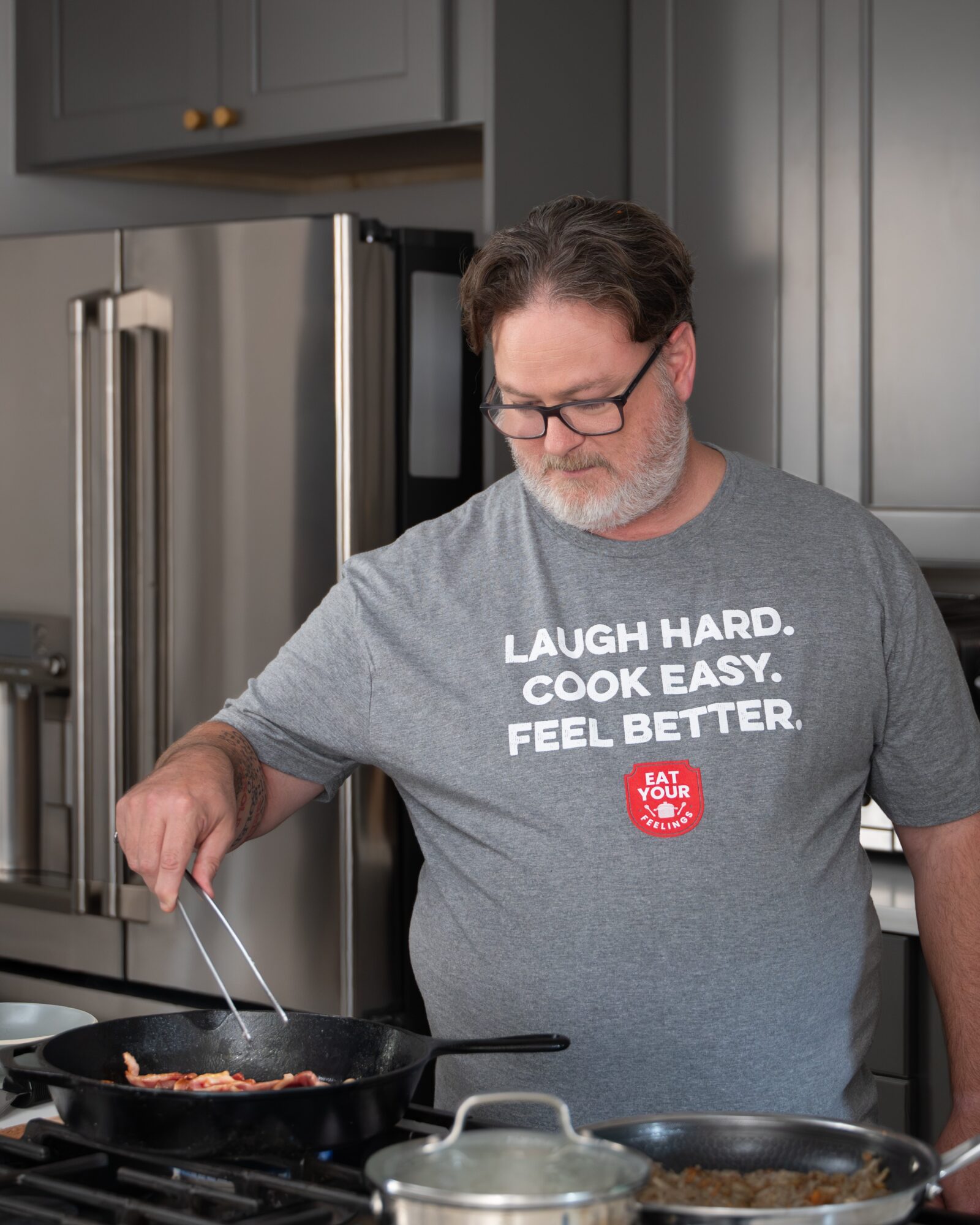 A person in a gray T-shirt stirs food in a frying pan on a kitchen stove.